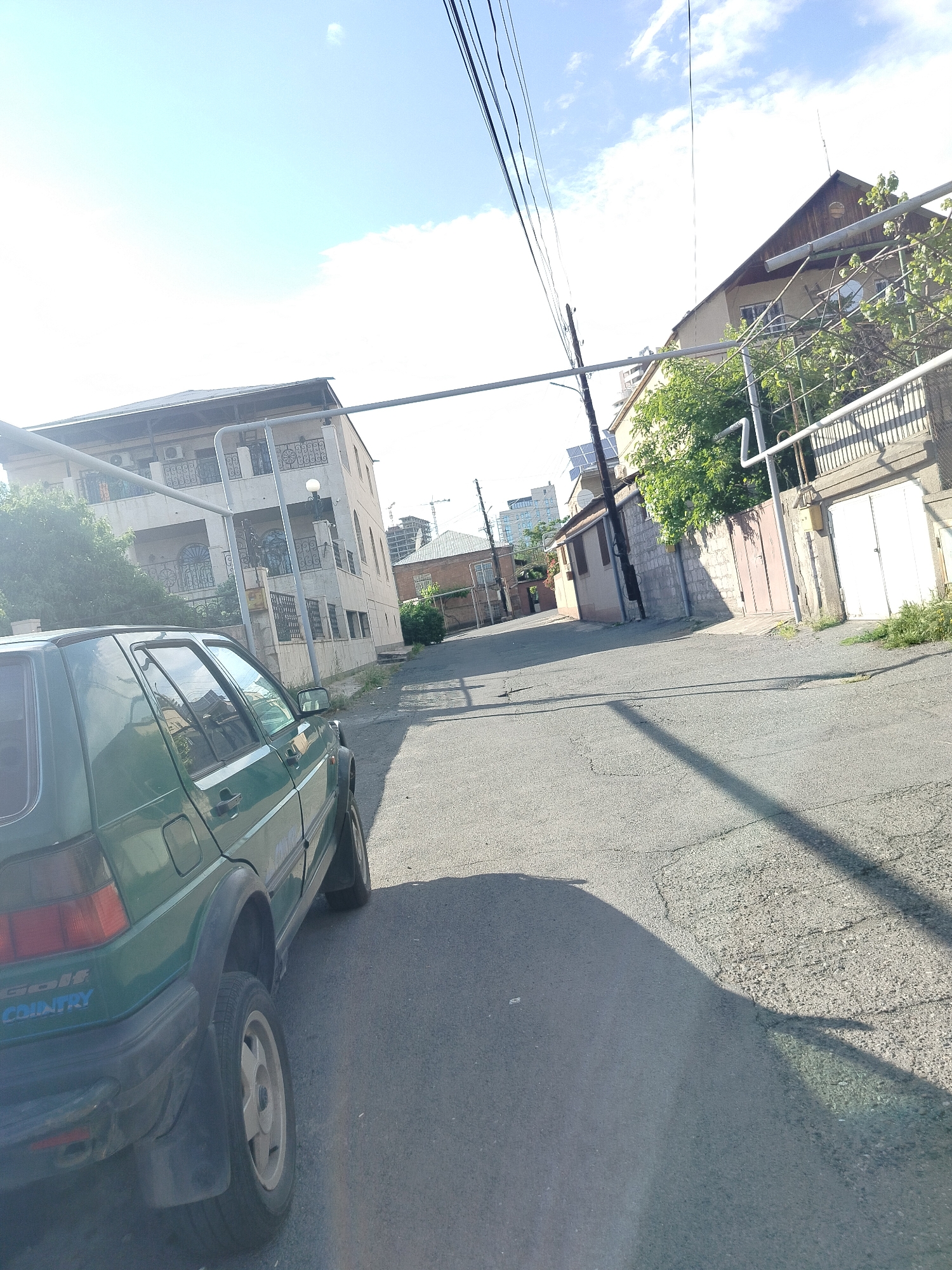 The image shows a residential street scene in Yerevan, Armenia. A green Volkswagen Golf Country is parked on the left side of the street. The street is paved with asphalt and shows signs of wear and repair. The street slopes gently uphill into the distance. Several buildings line the street, varying in architectural style and age. Some buildings are multi-story with balconies and decorative ironwork. Power lines and supporting poles are visible overhead. The sky is light blue with scattered clouds. The lighting suggests it is daytime.