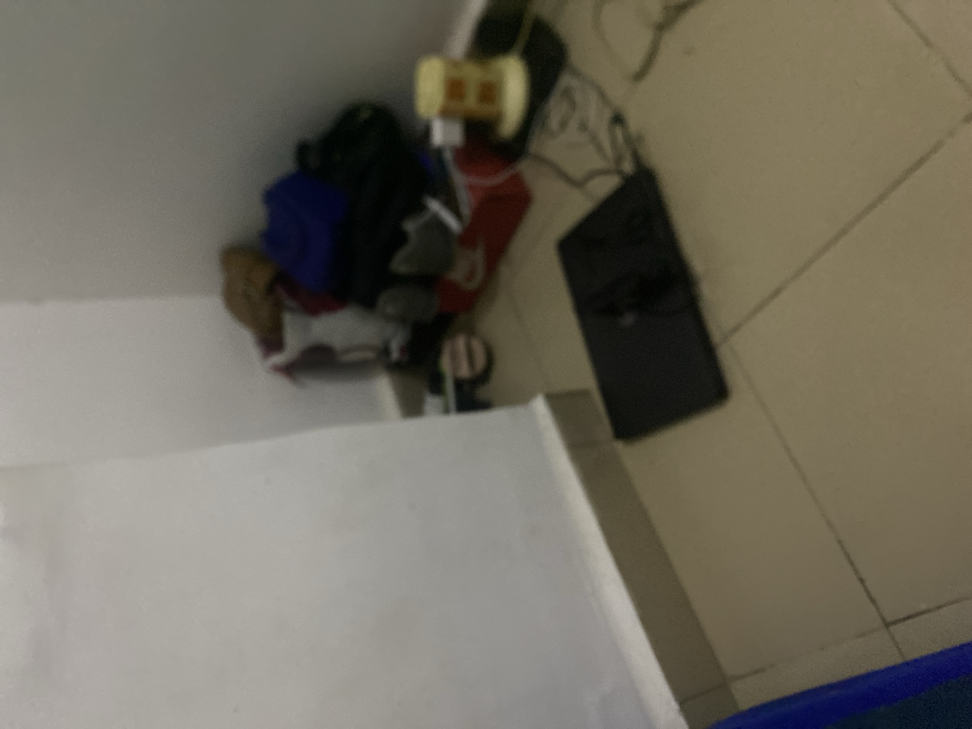 The image depicts a corner of a room, likely in an urban setting in Howayi, Nigeria. The walls are painted a light color and meet at a right angle. The floor is tiled in a neutral tone. In the corner, a pile of clothing and bags is stacked, suggesting a storage area or a messy corner. Among the items, a blue bag and a yellow object with a cylindrical shape and horizontal lines are visible. To the right of the pile, a red item, possibly a bag or box, is partially obscured.
In the foreground, a black laptop is placed on the floor, suggesting it is in use or temporarily set aside. Cables are visible near the laptop, connecting it to what appears to be a power source. The overall impression is of a functional, albeit somewhat disorganized, domestic space. The lighting in the room appears to be artificial, casting shadows and contributing to the slightly muted colors. There are no people visible in the frame.
