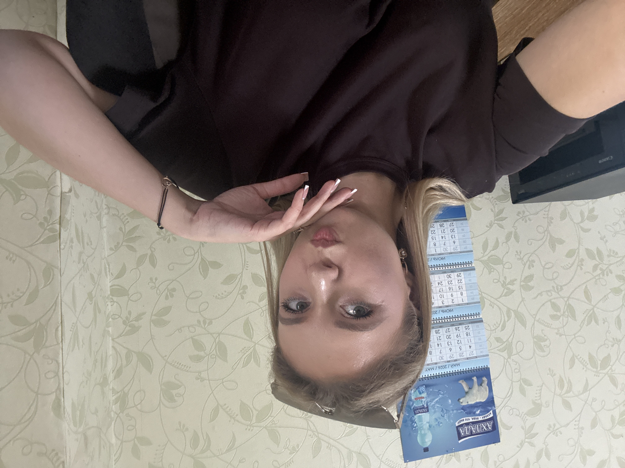 A young woman with blonde hair and makeup is taking a selfie. She is wearing a dark brown t-shirt and has sunglasses perched on her head. Her right hand is resting on her chin and her left arm is bent, with her hand supporting her head. She is wearing a black bracelet with a small heart charm. In the background, there is a calendar with images of a polar bear and a bottle of water. The wall behind her has a floral pattern. To the left of the frame, a black Canon printer is visible. The woman is making a kissing face.