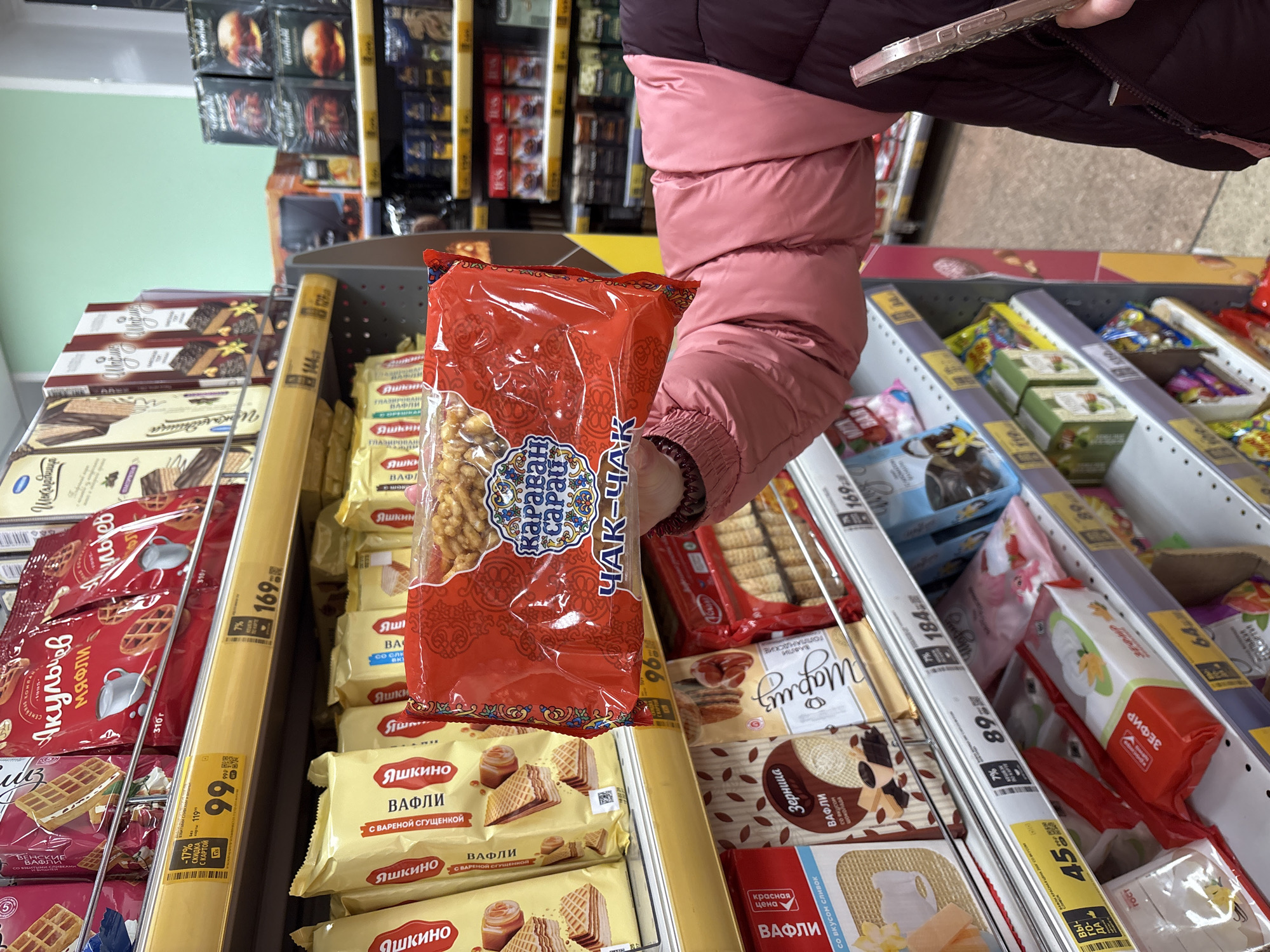 A person in a pink winter jacket holds a red package of "Karavan Sarai Chak-Chak" snack food in a store in Chaikovsky, Russia. The image focuses on the snack held up in front of shelves stocked with various wafer and biscuit products. The shelves are labeled with price tags displaying prices in Russian rubles, and the products include brands such as "Яшкино" (Yashkino) wafers, "Акульчев" (Akulchev) waffles, and "Зерница" (Zernitsa) wafers. The shelves are organized by product type with dividers and display individual prices. The person holding the snack is slightly out of focus, while the red package is more clearly visible. The background shows additional shelves of products, indicating a typical grocery store setting.