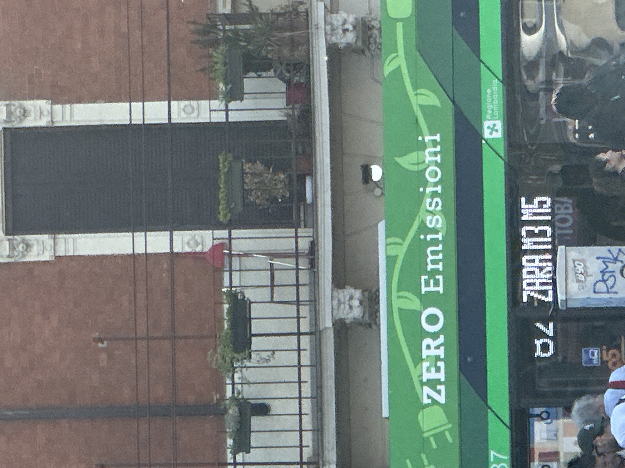 The image shows an outdoor scene in Milan, Italy. A passing "Zero Emissioni" bus with display "ZARA M3 M5" is prominent in the right foreground. A brick building with dark blinds, white trim, and wrought-iron balconies with plants is in the background. People are partially visible in front of the bus. A "Regione Lombardia" logo is visible on the bus, indicating the bus is operating in the Lombardy region of Italy.