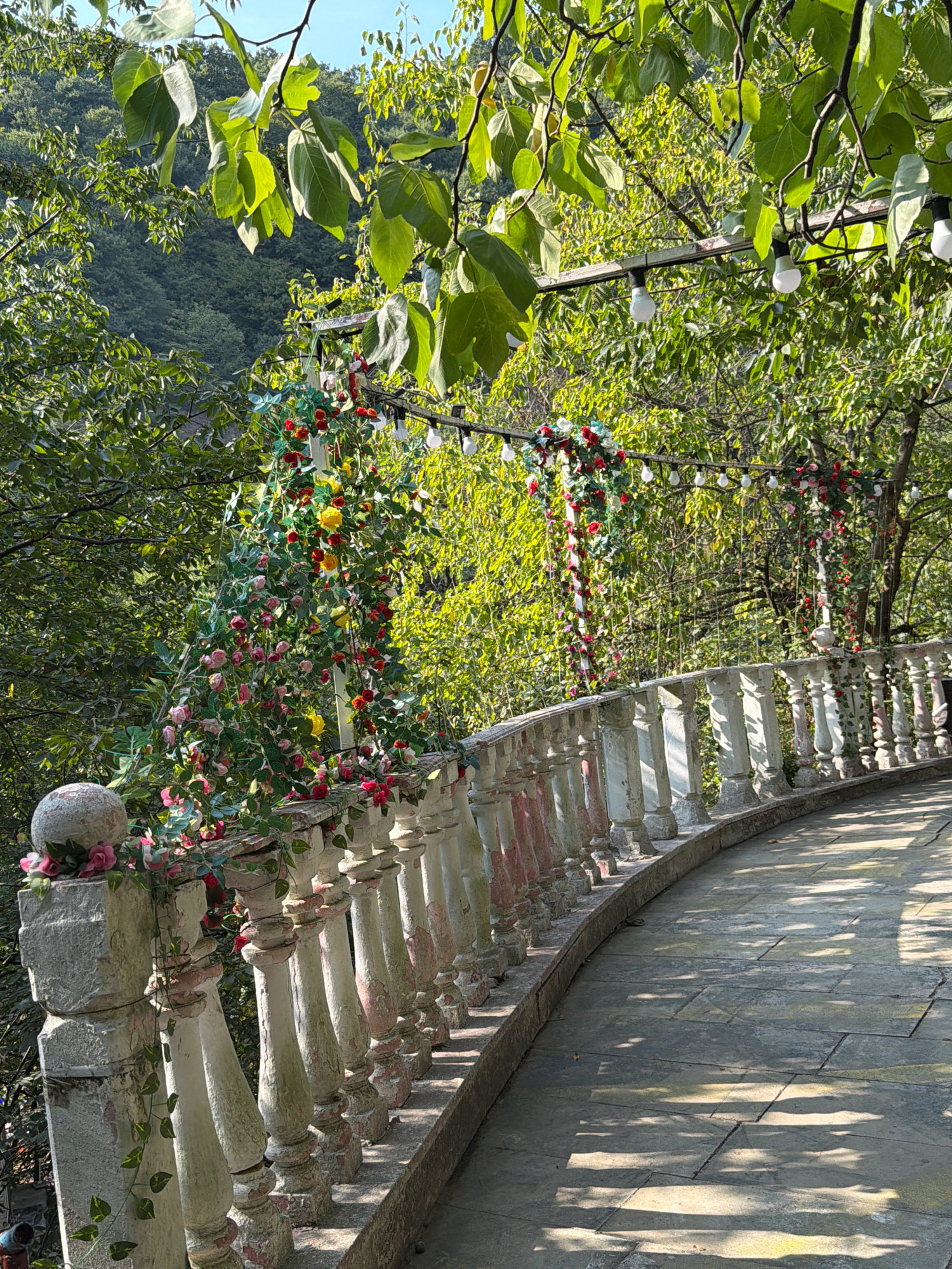 The image depicts an outdoor scene with a paved walkway curving to the right, bordered by a weathered stone balustrade. The balustrade is adorned with artificial flowers and ivy, with a spherical decorative element on the left post. Overhead, string lights are suspended from a wooden structure, interwoven with more artificial floral arrangements. Lush green trees and foliage dominate the background, suggesting a natural or park-like setting. The lighting indicates it is daytime, with dappled sunlight casting shadows on the walkway. No people are visible. The scene appears to be a decorated outdoor area, possibly for an event.