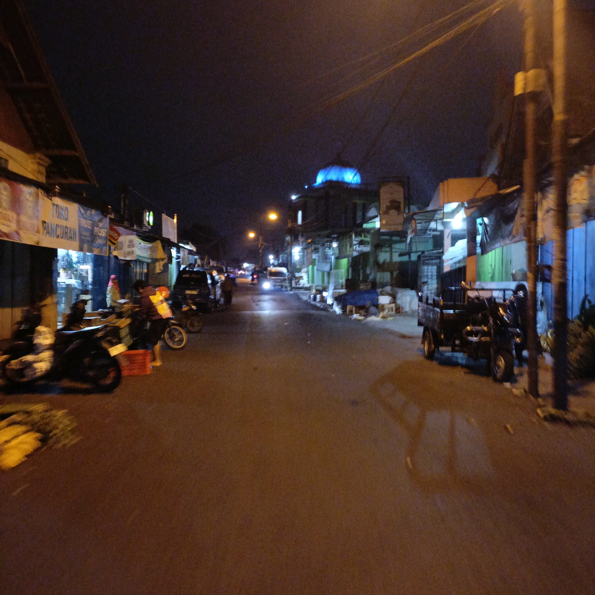 The image depicts a nighttime street scene in Salatiga, Indonesia, characterized by a busy, commercial atmosphere. The road is paved and shows some signs of wear. On the left side of the street, there is a row of shops with awnings and signage, including "TOKO PANCURAN." Several motorcycles are parked in front of these shops, some with their headlights on. One motorcycle is in the foreground, with its rear wheel blurred from motion or a long exposure. A person in a red headscarf stands near a stack of red crates. Further down the street, a white van with its headlights illuminated is driving away from the camera.
On the right side of the street, there are more shops and buildings. A prominent blue dome structure is visible in the distance, illuminated with blue light. A dark, three-wheeled cargo vehicle is parked on the right side of the road. A person is seen standing next to it. The street is lined with utility poles and wires overhead. The lighting is primarily from streetlights, shop lights, and vehicle headlights, creating a somewhat grainy and atmospheric night scene. The overall impression is of a lively, working street in a city at night.