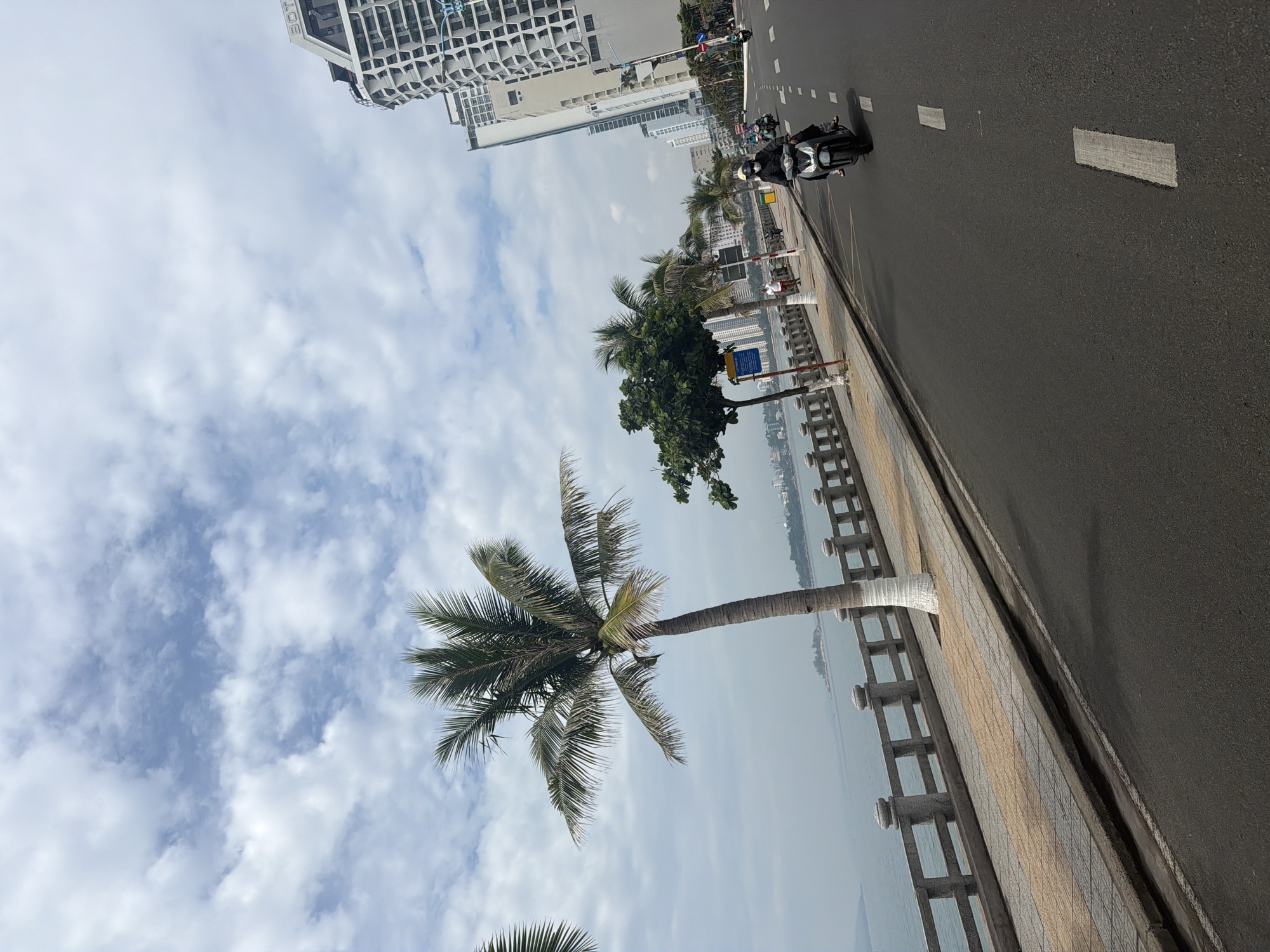 Captured from Bac Nha Trang Ward, Vietnam, the image shows a street scene on a cloudy day. A palm tree stands near a tiled walkway bordered by a gray balustrade facing the water. The road, marked with white lines, accommodates a scooter. High-rise buildings are visible in the background.
