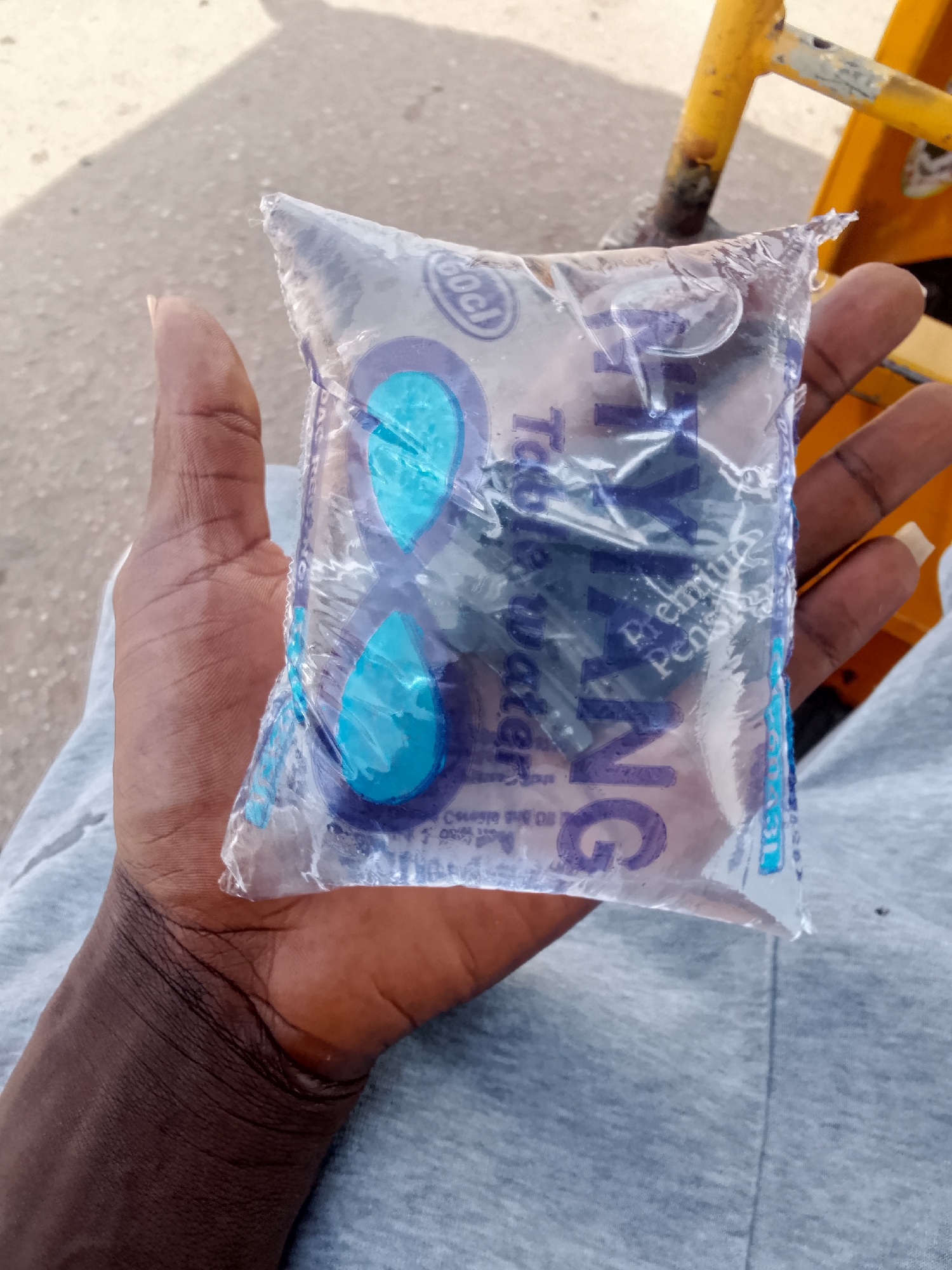 A close-up shows a dark-skinned hand holding a clear plastic sachet of water. The water sachet has blue and purple designs and lettering visible through the water. The hand is resting against a light gray fabric surface. Parts of a yellow metal frame or structure are visible in the background. The image was taken in Bukuru, Nigeria.