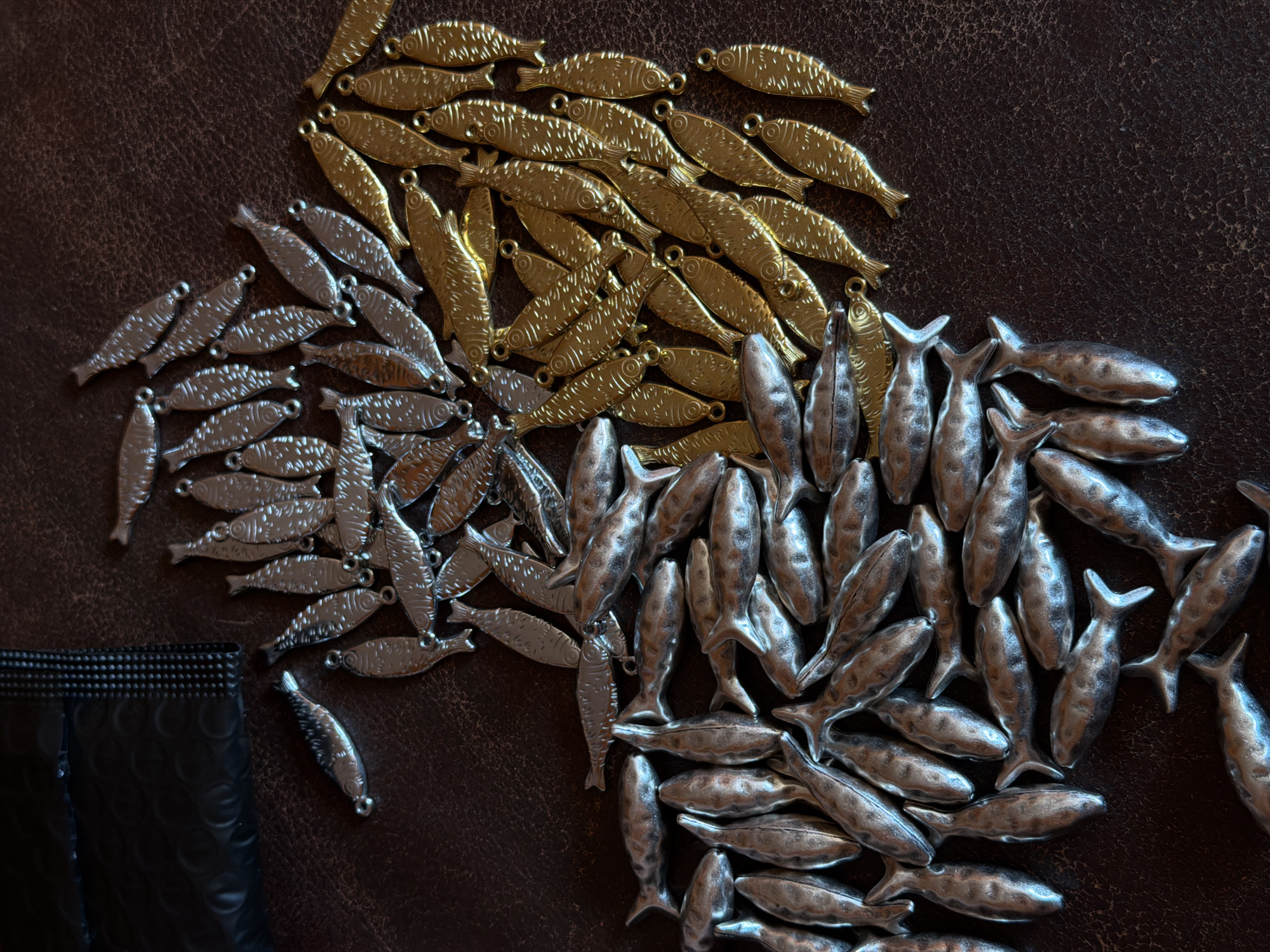 The image shows a scattering of metal fish charms on a textured, dark brown surface resembling leather. The charms are of three distinct types: gold-colored charms with detailed scale patterns, silver-colored charms with similar scale patterns, and larger, silver-colored charms with a more three-dimensional, sculpted appearance. The gold charms are grouped together at the top right, the patterned silver charms are located on the left side, and the larger silver charms are arranged at the bottom right. A small portion of a black, textured package is visible in the lower left corner. No people, subjects, or actions are visible. There is no indication of this image being located in Khimki, Russia.