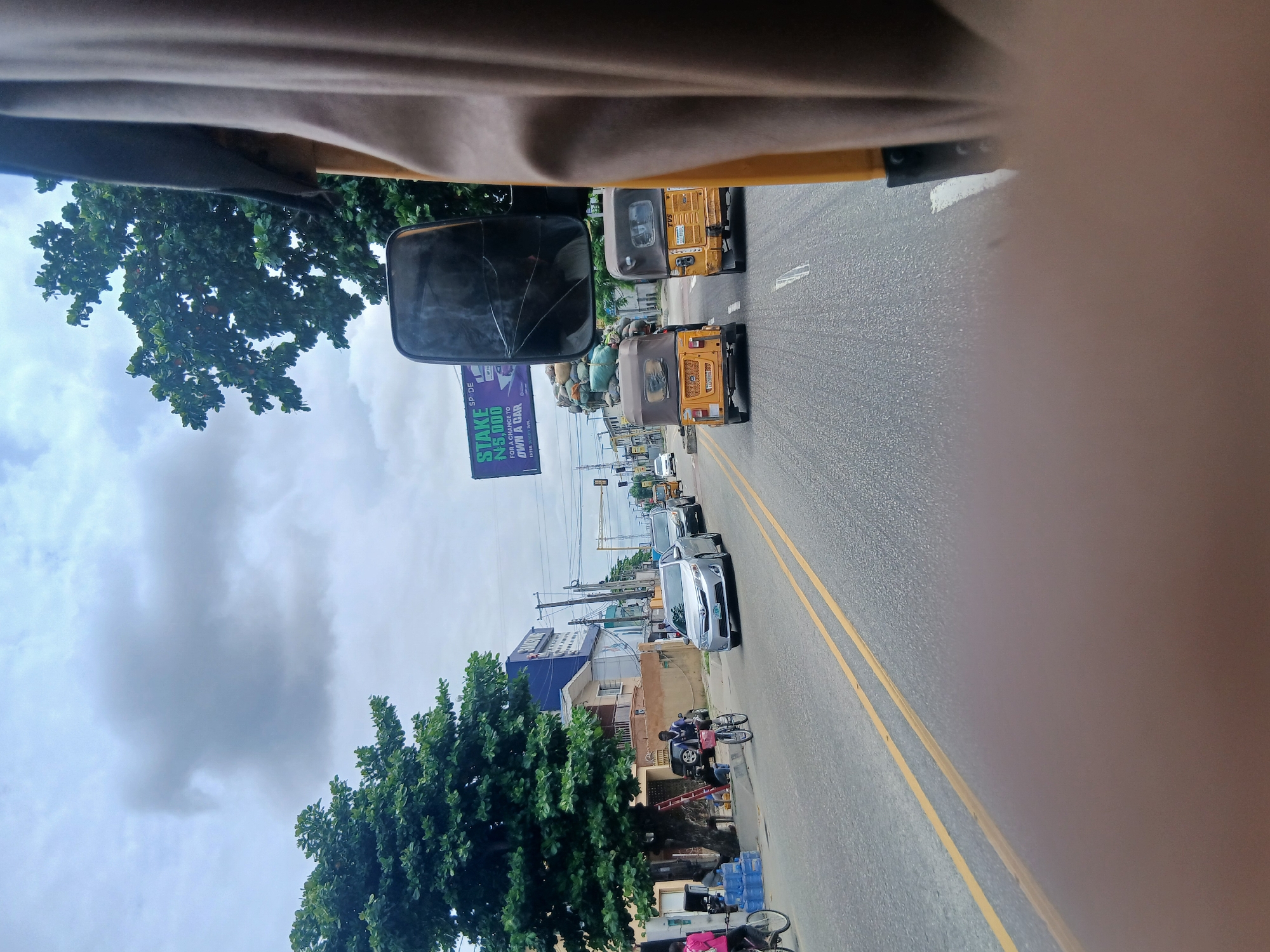 The image captures a street scene in Animashawun, Nigeria. The perspective is from inside a vehicle, looking forward and slightly to the side. The foreground shows parts of the vehicle's interior, including what appears to be a dashboard and a large, cracked side mirror.
The street is paved and marked with double yellow lines. Several yellow auto-rickshaws, also known as 'keke napep', are visible in the middle and right lanes, suggesting they are part of the local transportation system. A silver car is in the right lane ahead of the keke napeps. The left side of the street features buildings and dense green trees. A large billboard is also visible on the left, with text including "STAKE N5,000 FOR A CHANCE TO OWN A CAR".
Further down the road, there are more buildings, traffic lights, and utility poles, indicating a moderately developed urban or suburban area. People are seen on the side of the road, with one person on a bicycle and another near a parked vehicle. Some blue water bottles are stacked on the sidewalk. The sky is overcast with large, grey clouds, suggesting it might be a cloudy or potentially rainy day. The overall impression is of a busy street with everyday activity.