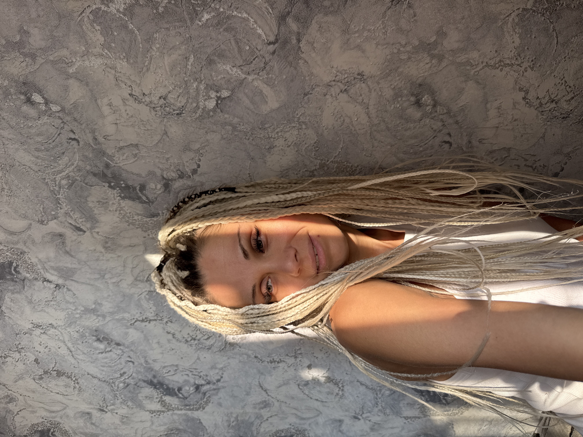 A young woman with long blonde braids is leaning against a textured gray wall in Rusanovo, Russia. Sunlight is casting shadows across her face and shoulder. She is wearing a white tank top. The wall has a marbled pattern with variations in gray tones.