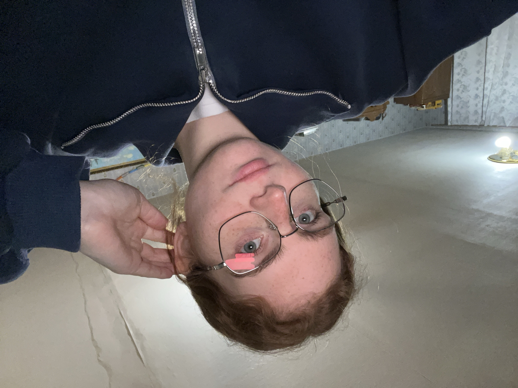 The media file features a selfie of a person with fair skin, light-colored (blue/grey) eyes, and reddish-brown hair. They are wearing silver-rimmed glasses and a dark blue zippered hoodie over a white undershirt. A hand is visible near their right ear. The image is captured from a low angle, looking up at the subject, who appears inverted in the frame, directly engaging the viewer with a neutral expression. A pink censor bar is placed over the subject's left eye.
The setting is an indoor space, likely a basement or an older room, located in Новая Тура, Russia. The background includes a light-colored, somewhat reflective floor. One wall is partially covered with patterned wallpaper (a light, possibly floral or abstract design on a pale background), while an adjacent section shows significant wear, with wallpaper torn away to expose wooden structures beneath.
A notable object in the background is a bare light bulb, resting on a decorative, possibly metallic, base, emitting a bright artificial light. This suggests the image was taken indoors, likely at night or in a poorly lit area, relying solely on this artificial light source. The overall impression is a candid moment captured in a utilitarian or older residential interior, possibly an area undergoing renovation or in a state of disrepair, indicated by the exposed structural elements and simple lighting. There is no visible text in the image.