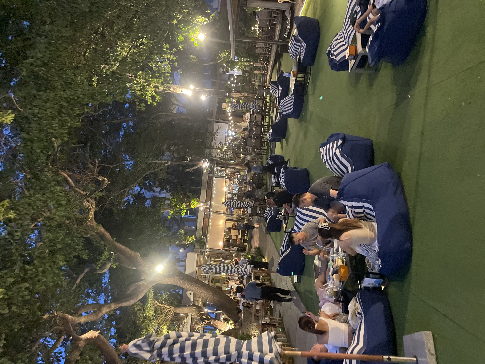 The image depicts an outdoor dining area at night in Pattaya City, Thailand. Patrons are seated at low tables on beanbag chairs and traditional seating arrangements, enjoying food and drinks. The ambiance is enhanced by string lights and overhead lamps. The scene is set amidst lush trees, providing a natural canopy. The overall atmosphere suggests a relaxed evening gathering at an establishment that offers both comfortable lounging and dining experiences.