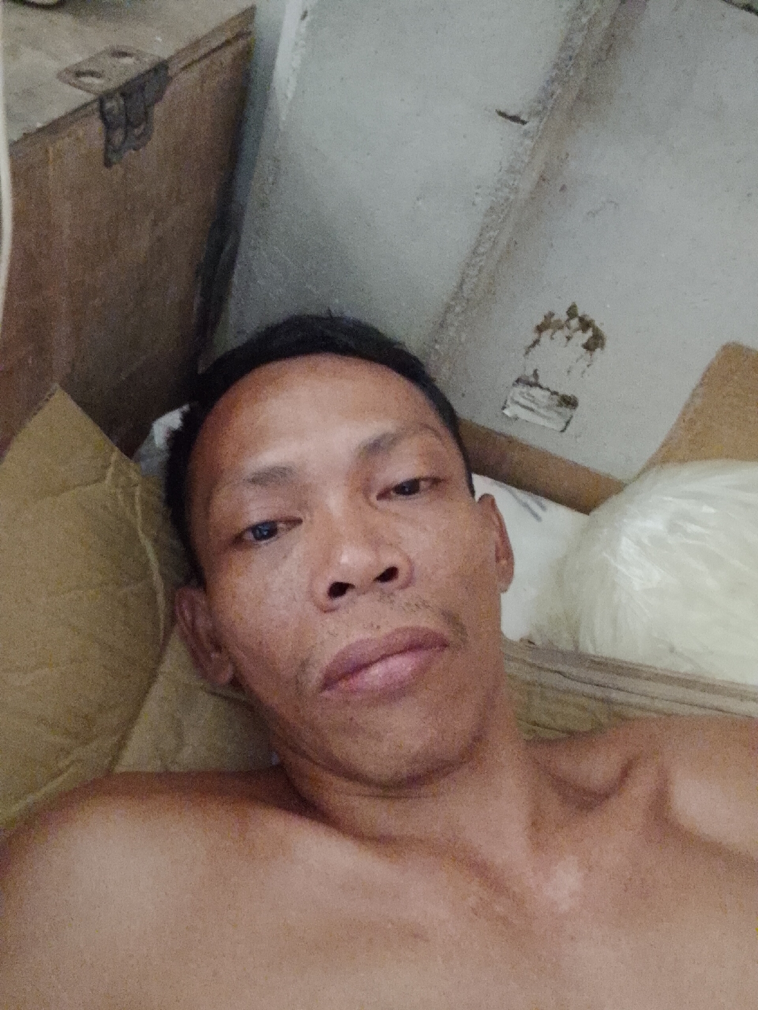 An adult male with dark hair and skin is visible in a close-up, front-facing shot. He appears to be lying down, as suggested by the angle and the visible portion of his bare chest and shoulders. The setting is a cluttered space with a wooden box and a concrete wall behind him. The location is Bukit Ketok, Indonesia.