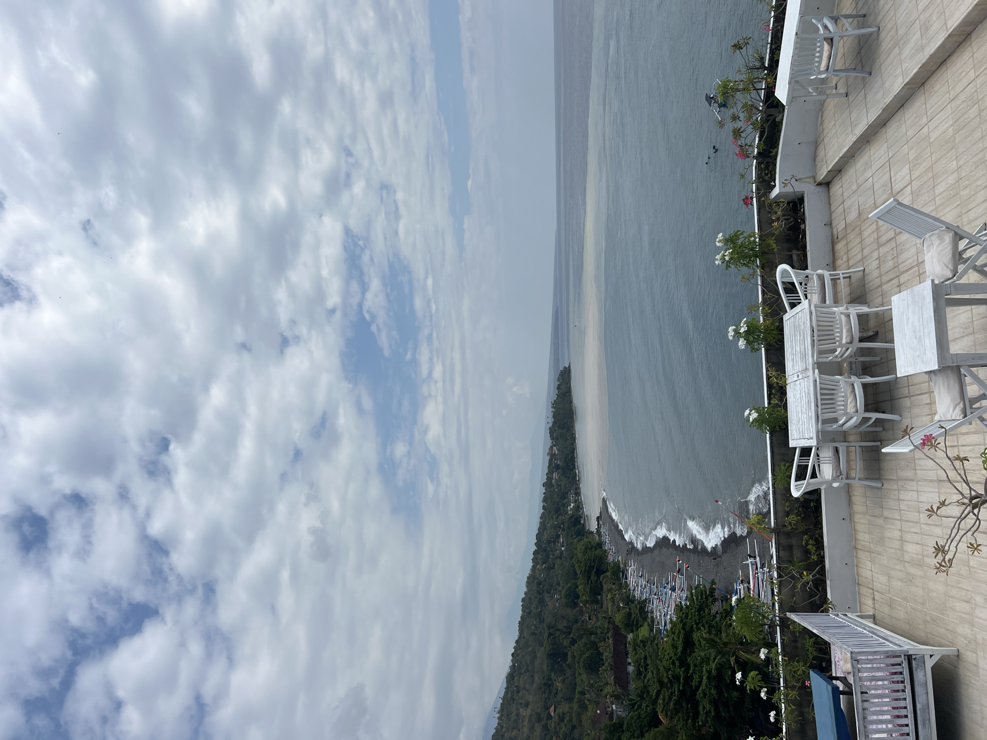 An elevated, medium shot captures a coastal view from a rooftop patio in Abang, Indonesia. The patio features white wooden tables and chairs, some set for dining, others arranged for lounging. A white-railed bench with a blue cushion occupies the left side of the foreground. Beyond the patio, a dark, pebbled beach is visible, lined with numerous traditional outrigger fishing boats. The sea, a gradient of deep blue to lighter, sandy hues near the shore, stretches to the horizon under a cloudy sky. Lush green hills, dotted with buildings, rise behind the beach, forming a distant coastline. The overall scene depicts a tranquil seaside resort or restaurant.