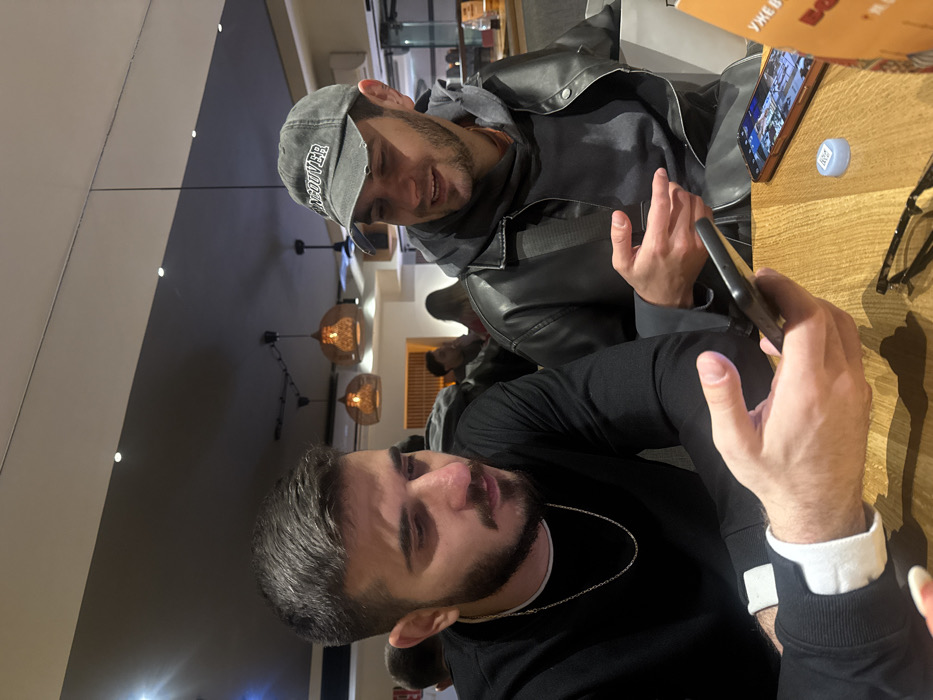 Two men are seated at a table, engrossed in a mobile phone. The first man is wearing a black long-sleeved shirt with a silver chain necklace. The second man is clad in a gray hooded sweatshirt and a black leather jacket, also looking at the phone. A second phone is lying on the table along with a water bottle cap and a pair of eyeglasses. In the background, other people are visible, and decorative light fixtures illuminate the scene.