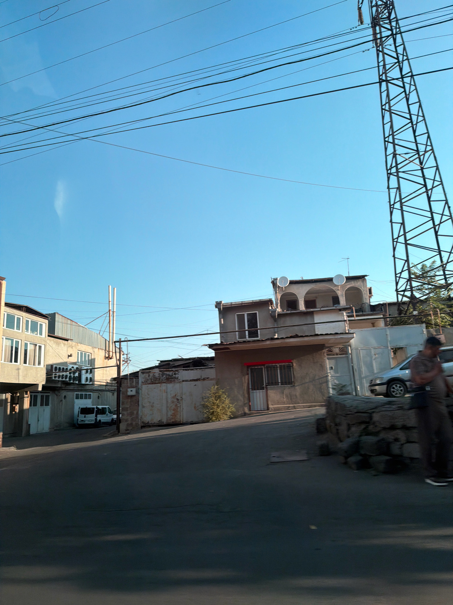 The image depicts a street scene in Yerevan, Armenia, characterized by residential buildings, overhead power lines, and a prominent electrical pylon. The buildings appear to be a mix of older and newer constructions, with some featuring balconies and satellite dishes. A white van is parked in front of one of the buildings. A person, partially obscured, is standing near a silver car on the right side of the image. The sky is clear and blue, suggesting daytime. The road in the foreground is asphalted, with some shadows cast across it.