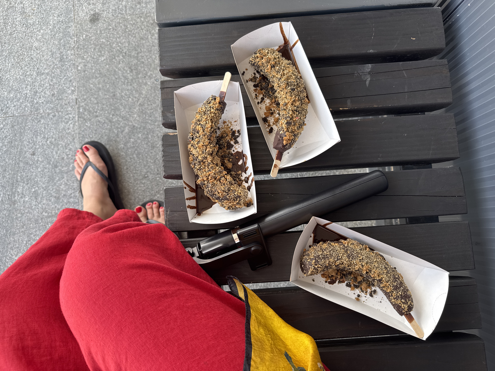 In Žanjev Do, Montenegro, a person sits on a dark wooden bench, their legs covered in vibrant red trousers. One foot, adorned with red nail polish, is visible in a dark flip-flop, resting on a grey, textured surface. In front of the person, three chocolate-dipped bananas, coated in a crumbly topping and served in white paper trays, are arranged on the bench. A black object, possibly a camera or a small bag, lies next to the person's leg, partially obscured by a yellow and black patterned fabric. The overall scene suggests a casual outdoor setting, likely a park or a public seating area.