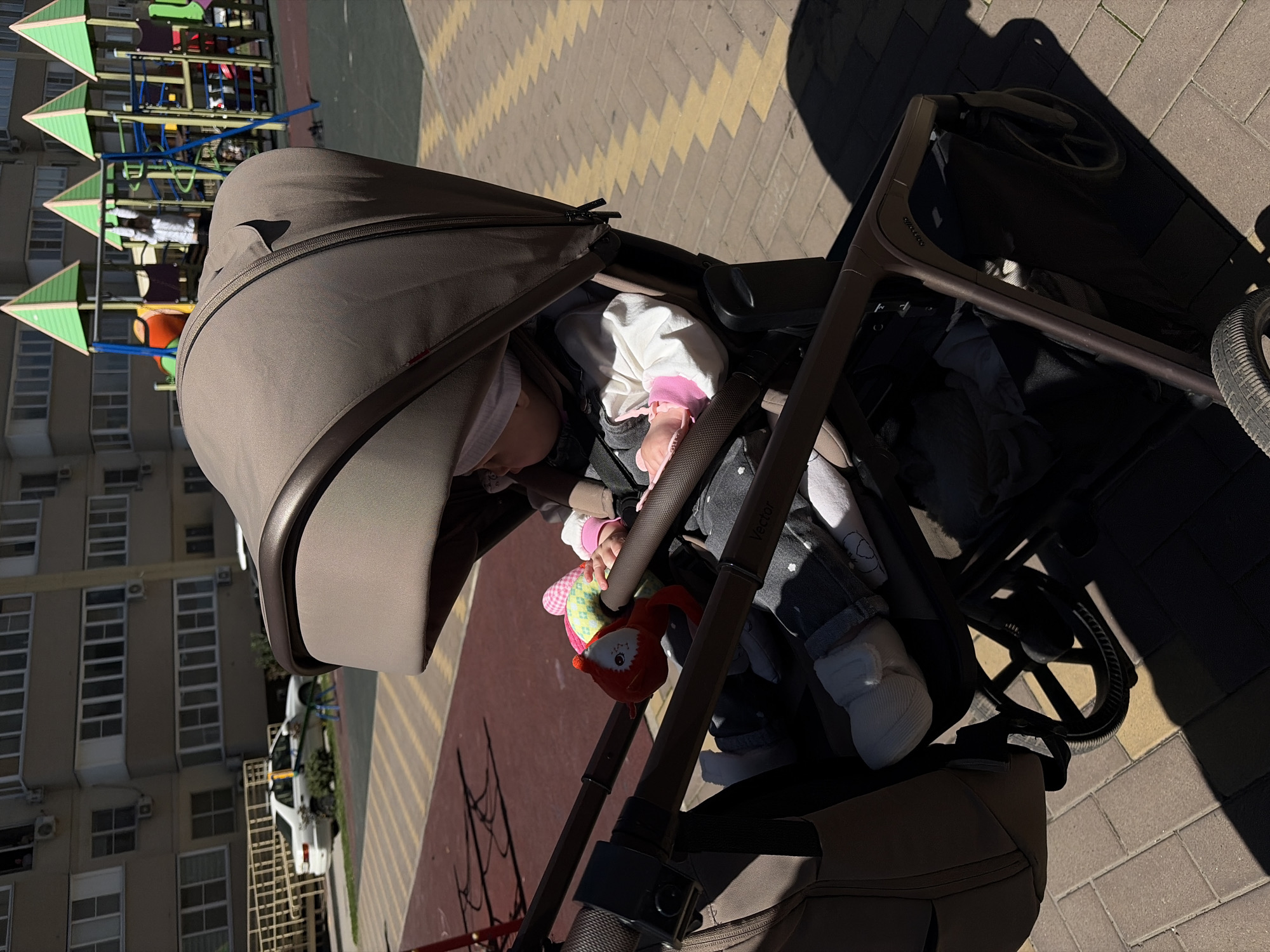 A baby is sleeping in a stroller on a brick sidewalk in what appears to be Krasnodar, Russia. The stroller is brown, with a large sunshade covering the baby. The baby is wearing white and gray clothing with a pink detail, and a white hat. A soft toy, possibly a fox, hangs from the stroller's safety bar. In the background, there is a building with many windows, as well as a playground. The area is paved with light-colored bricks. The scene is well-lit, suggesting it's daytime.
