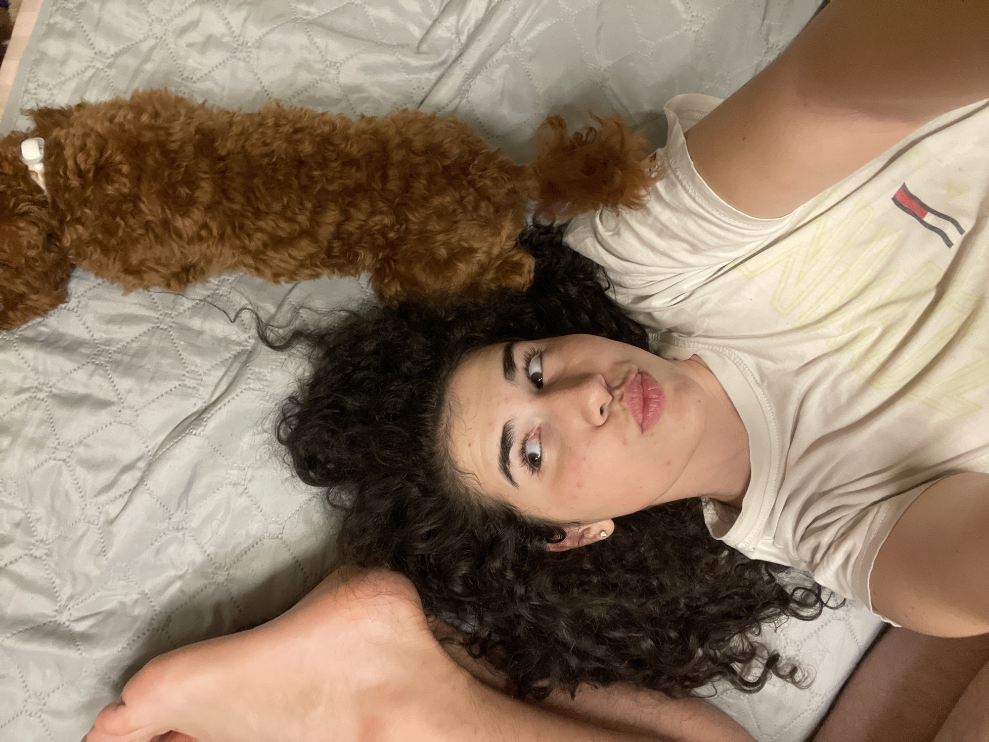 A high-angle, close-up shot shows a young woman with dark, curly hair lying on a light blue, quilted bedspread. Her head is tilted to the right, and her eyes are looking upwards and to the right. She has her lips pursed, and a faint blush is visible on her cheeks. A small, brown, curly-haired dog is lying on the bedspread next to her head, with its body extending towards the top of the frame. The woman is wearing a light-colored t-shirt with a graphic design. A person's left foot is visible in the upper left quadrant of the image, positioned near the woman's head. The scene appears to be indoors, likely in a bedroom.
