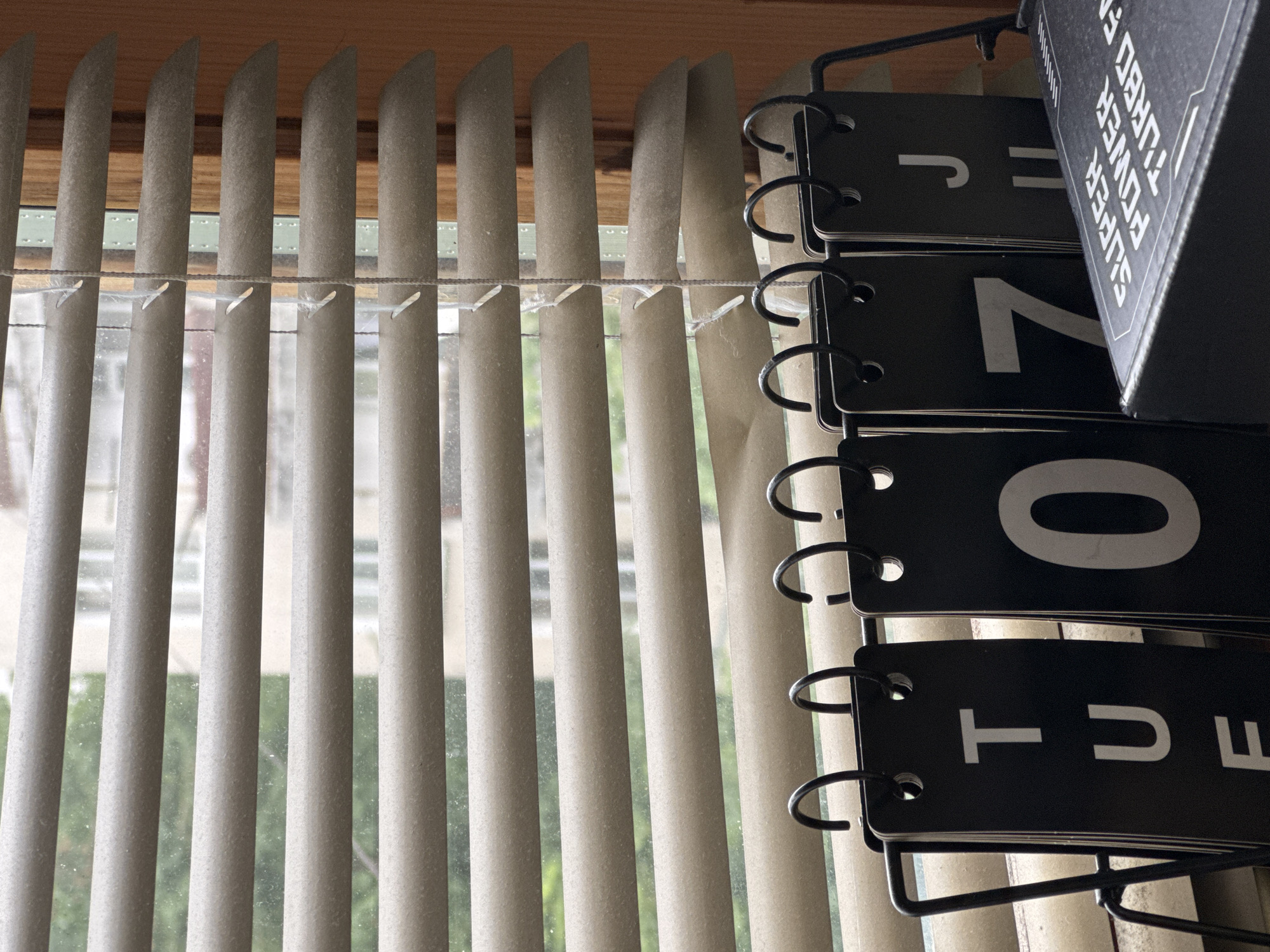 The image shows a close-up view of a window with beige horizontal blinds. Behind the blinds, a blurred outdoor scene with trees and buildings is visible. In the foreground, a black metal stand holds a flip-style calendar made of black cards with white lettering. The calendar displays "TUE" on the first card, followed by "0" and "7" on subsequent cards, and "J" on a card further to the right. A black box with white lettering, reading "SUPER POWER TURBO FA", is placed in front of the calendar. The overall scene appears to be indoors, likely in a room near a window. There are no people or animals present in the image. The lighting suggests it is daytime. The specific location in Smolyan, Bulgaria cannot be determined from the image content.