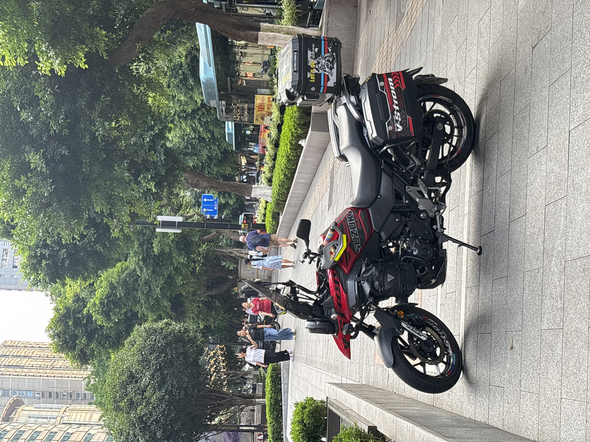 A horizontal shot captures a Suzuki V-Strom motorcycle parked on a gray tiled sidewalk in Yuzhong District, China. The motorcycle is red and black, with the "V-Strom" logo visible. A black storage case is mounted on the rear, adorned with stickers. The bike is positioned on its kickstand.
In the background, a group of people stroll along the sidewalk, with the greenery of trees and shrubs on the side. A street sign is also visible. Beyond the trees, a building with an arched roof can be seen.