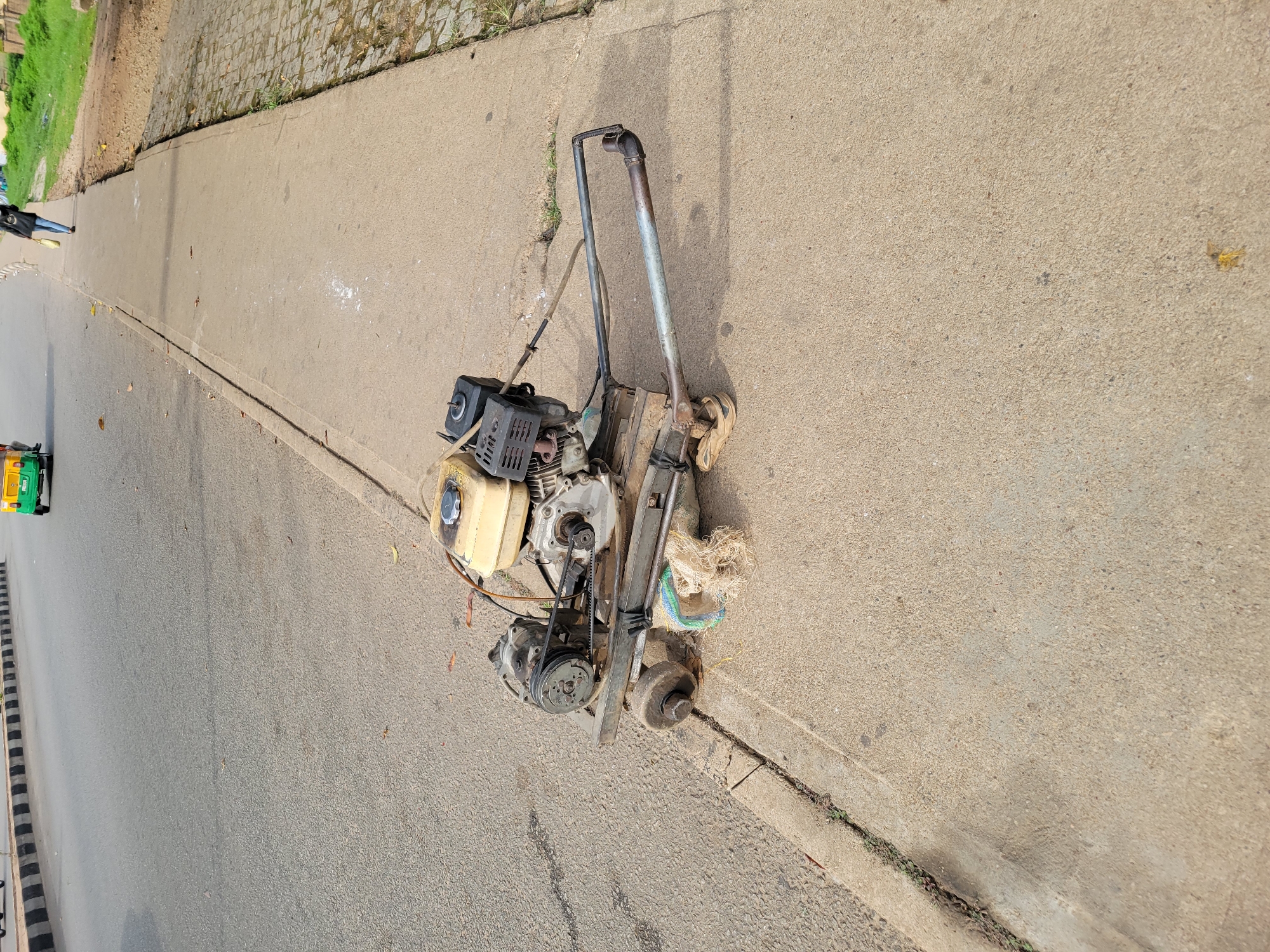 A gasoline-powered plate compactor is the central subject of the image, positioned on a light grey concrete sidewalk. The compactor features a visible engine with a light-colored fuel tank, an exposed belt-drive system, and metal handles extending upwards. A bundle of fibrous material, possibly netting or rags, is tucked near the machine's base. The sidewalk shows signs of wear and has some small white markings.
To the left of the sidewalk, a dark asphalt road runs parallel. A green and yellow auto-rickshaw is partially visible on the road in the lower left background, and further down the sidewalk, a person wearing dark clothing is walking. Beyond the sidewalk, there's a strip of dirt and patchy green grass, leading to a section of textured paving, possibly cobblestones.
The setting is an outdoor urban or semi-urban street, illuminated by clear daylight, casting discernible shadows. The compactor appears to be stationary and not in operation. Faint text, "HONDA," is visible on the engine's fuel tank area. A small yellow insect is present on the sidewalk surface in some detailed views.