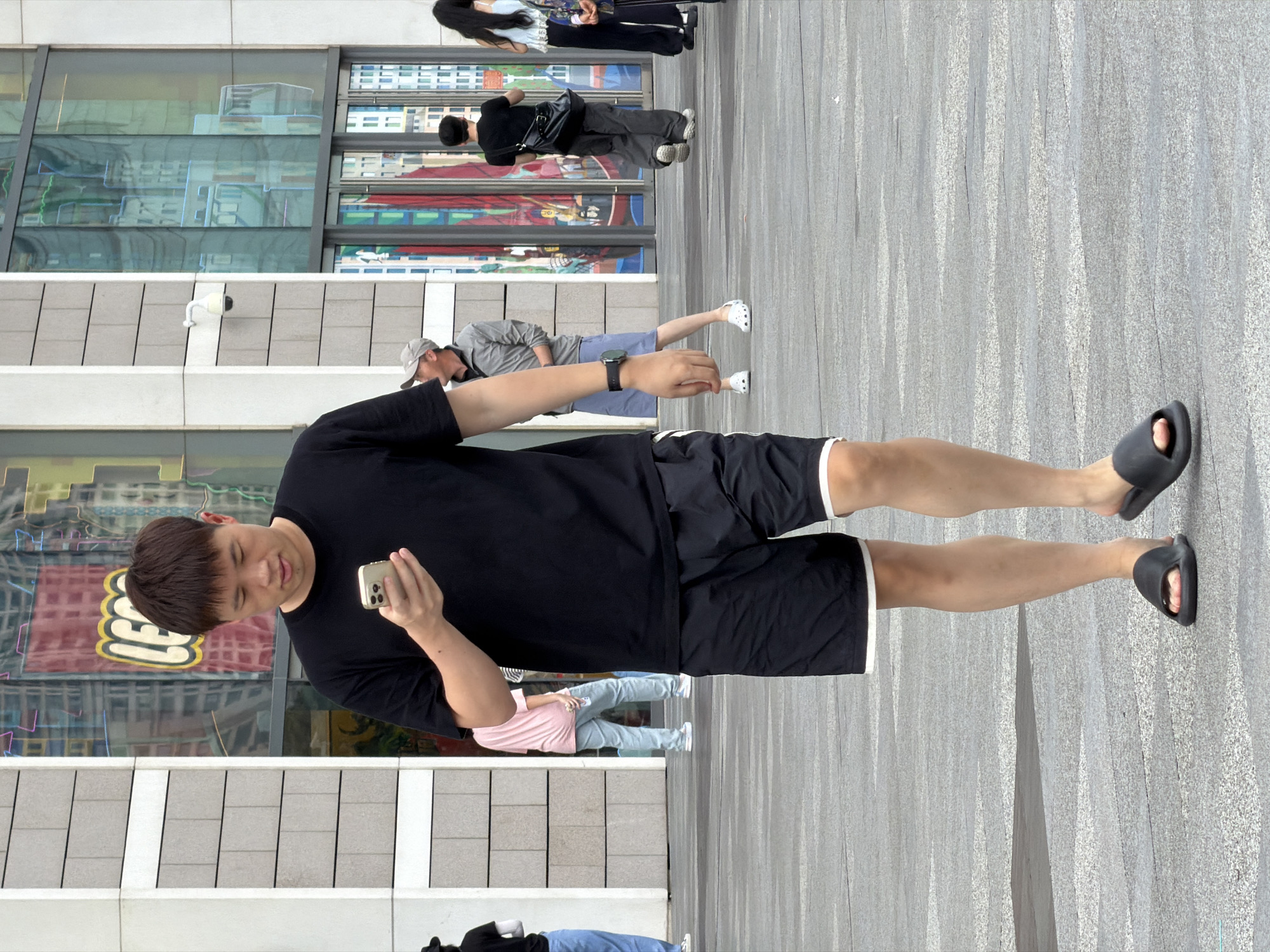 A young Asian man wearing a black t-shirt and black shorts with white trim walks on a grey paved outdoor area, holding a smartphone and looking at its screen. He is wearing black slides on his feet. In the background, a building with a facade of grey rectangular panels and large glass windows is visible. The windows reflect images of buildings and neon lights. A red and yellow LEGO logo is displayed on a banner visible through one of the windows. Other pedestrians are present in the background, including a person in a pink t-shirt and light blue jeans, and a person wearing a grey cap and jacket. The scene appears to be a public plaza in Yuzhong District, China.