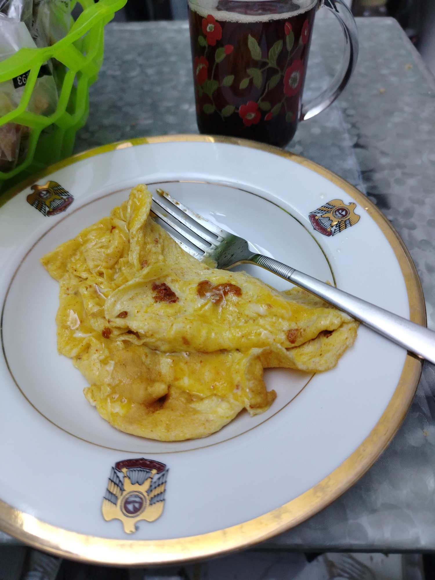A close-up shot shows a plate of scrambled eggs, partially eaten, with a fork lying across it. The plate is white with a gold rim and features several small, emblem-like designs spaced around the edge. A glass mug filled with a dark liquid and decorated with red floral patterns sits behind the plate. Part of a green basket is visible in the upper left corner. The scene appears to be set on a textured, slightly mottled surface, possibly a table. There is no indication of this image being from Al Mas'oudi, United Arab Emirates.