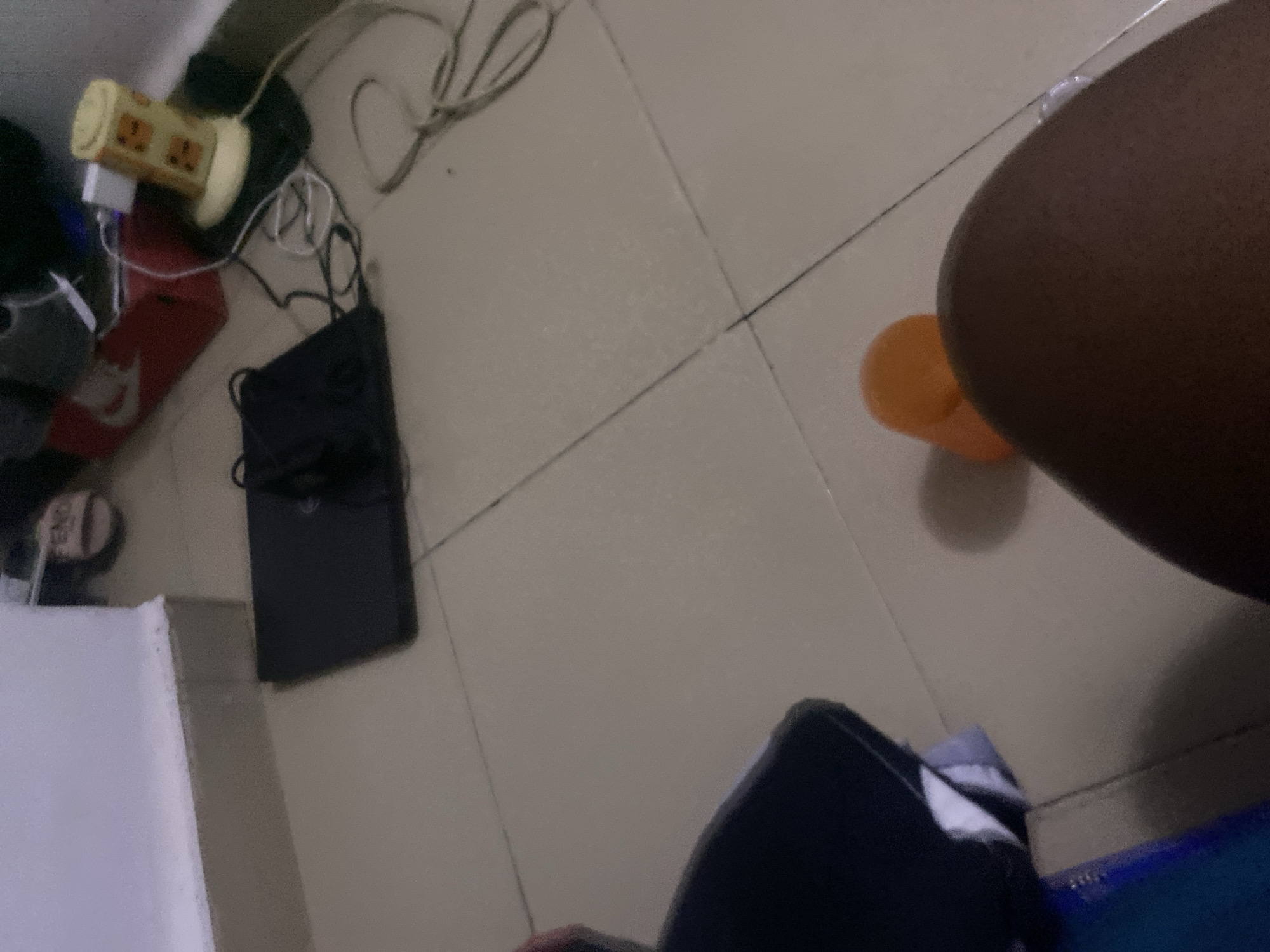 The image depicts a floor scene with tiled surfaces and a wall baseboard. On the floor, a closed black laptop is visible, with cables and a yellow desk lamp with a cartoon face pattern behind it. In the foreground, a person's leg and a small orange object, possibly a cup or toy, are in focus. A pair of pink slippers with the "FENDI" logo are also present. The environment appears to be an indoor setting, likely a room, with a neutral color palette. The lighting suggests it is indoors during the day or under artificial light. The scene does not provide specific visual cues to pinpoint a location within the city of Howayi, Nigeria.