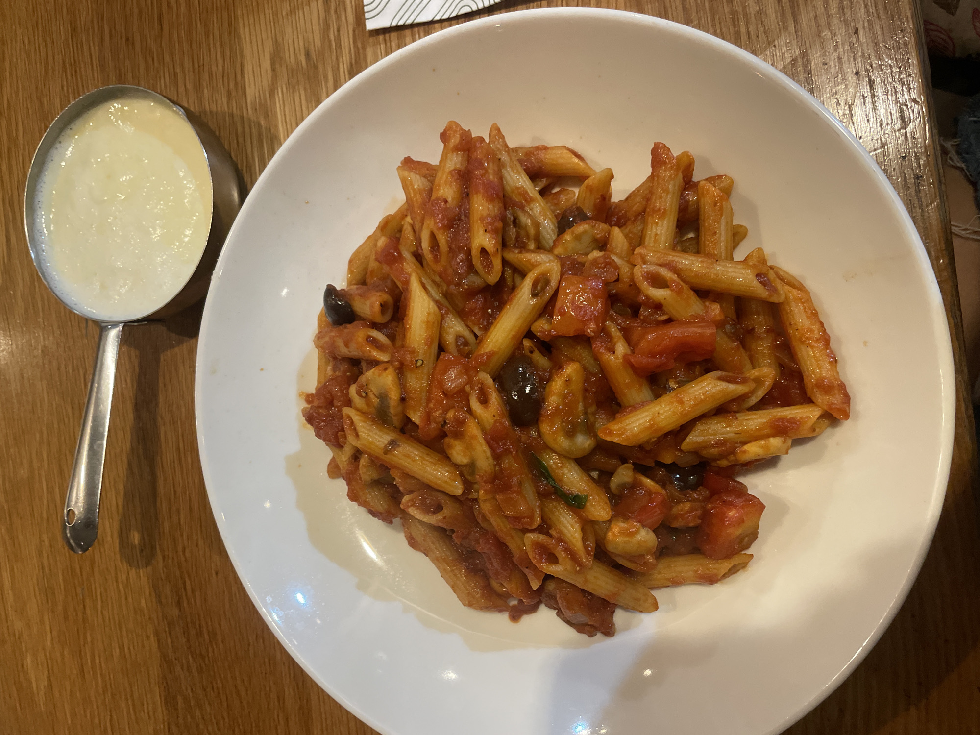 A close-up, overhead shot captures a white, shallow bowl filled with penne pasta coated in a chunky red sauce and what appear to be black olives and mushrooms, positioned on a wooden table. To the upper right of the bowl, a small, metallic pot containing a creamy, pale yellow sauce is visible. A metallic utensil with a hole in its end rests partially on the table, above and to the left of the bowl. The table's surface exhibits a wood grain pattern. This scene is located in Yokneam Ilit, Israel.