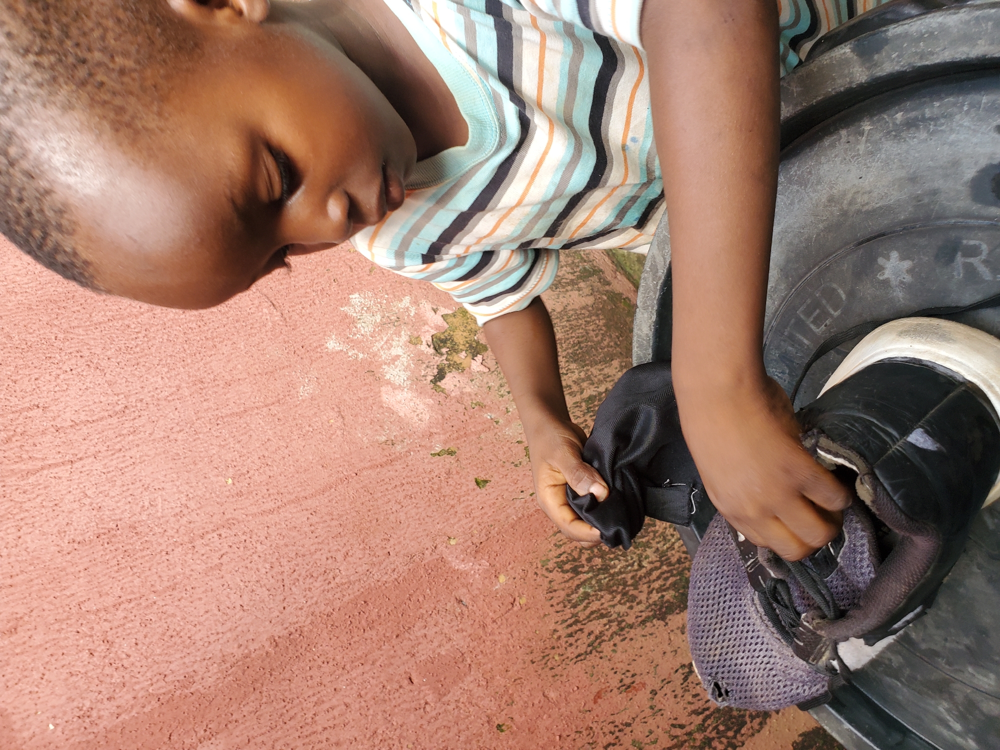 A young child, appearing to be around 4-5 years old with dark skin and a shaved head, is focused on a pair of black and white sneakers resting inside a large, dark plastic basin. The child wears a horizontally striped t-shirt with blue, orange, and black bands. Their hands are engaged with the shoes, one holding a black cloth or bag, the other appearing to be manipulating the shoe's laces or interior. The background features a textured, salmon-pink wall, typical of some outdoor urban environments in Jos, Nigeria, with some areas of peeling paint and moss growth, suggesting the setting is likely an outdoor area of a residence or public space. The lighting suggests it is daytime, casting soft shadows.