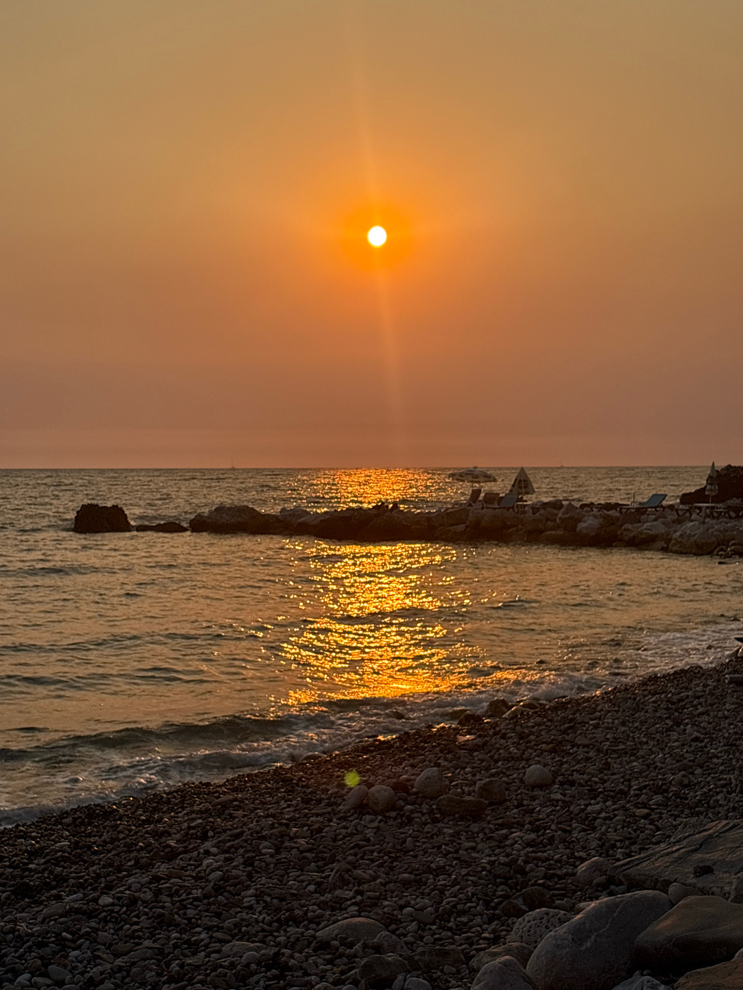 This is a photograph of a sunset over the Adriatic Sea in Bar, Montenegro. The sun is a bright orange orb low in the sky, casting a golden glow on the water and the beach. The water is calm, with gentle waves lapping at the shore. The beach is covered in dark gray and black pebbles, with larger stones scattered throughout. In the distance, there is a rocky outcropping with some lounge chairs and umbrellas. The sky is a hazy orange, with the sun's rays visible as shafts of light. A small sailboat can be seen on the horizon. The overall mood is peaceful and serene.