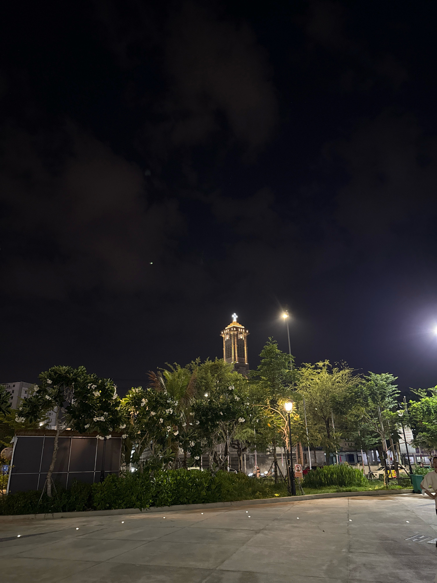 The image captures a night scene in Nha Trang, Vietnam, focusing on a park-like area in front of a prominent church tower. The sky is dark with scattered clouds, and a faint celestial body is visible. The church tower, illuminated by internal lighting and a cross at its apex, stands tall, surrounded by lush green trees. Streetlights illuminate the pathway and surrounding vegetation, casting a warm glow. The foreground shows a paved walkway with small embedded lights. The scene suggests a tranquil evening in a public space, possibly a church plaza or a park adjacent to a religious establishment.