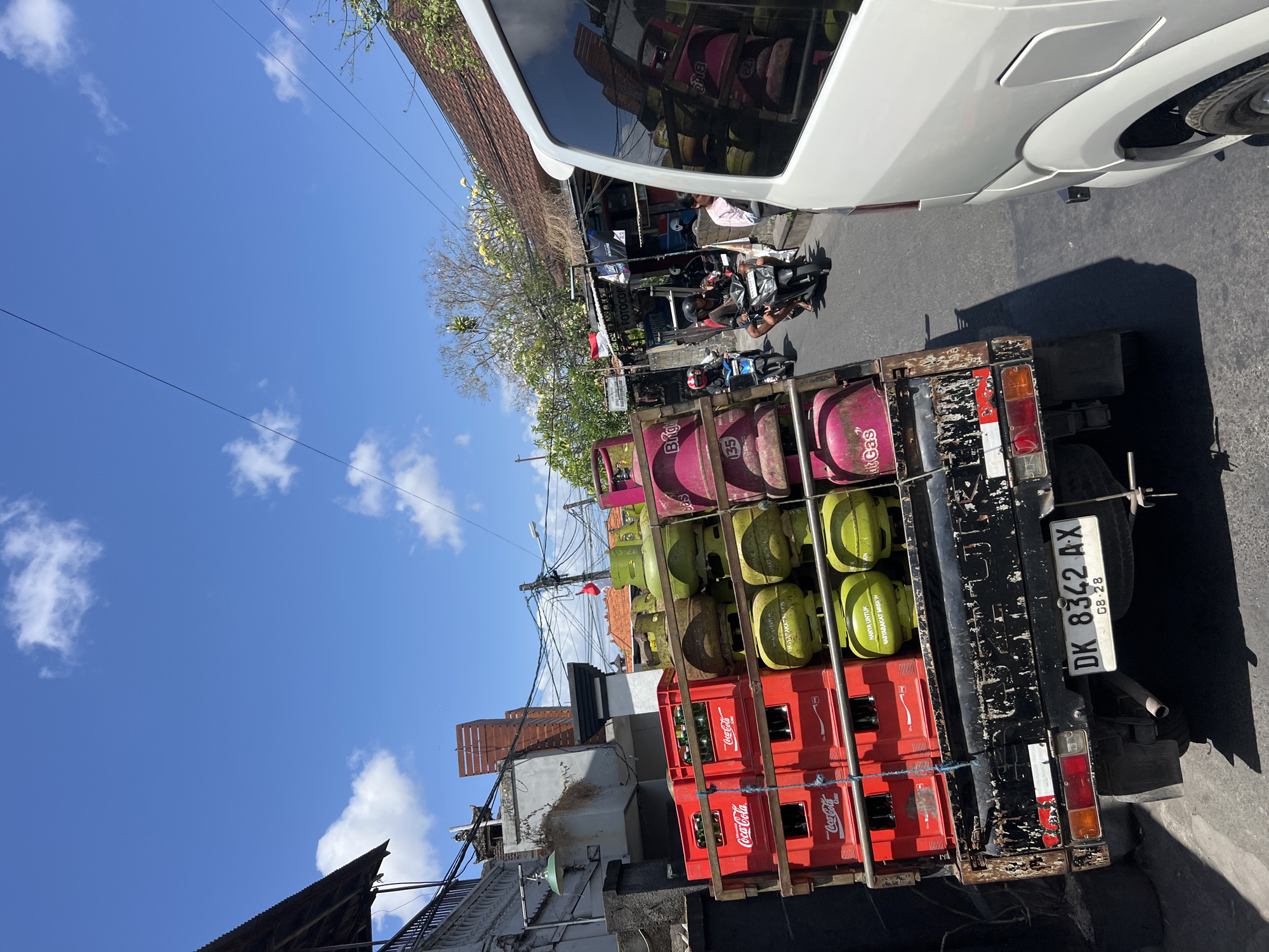 This image captures a street scene in Denpasar, Indonesia, on a sunny day. A truck, overloaded with green and pink gas cylinders and red Coca-Cola crates, is positioned on the right side of the frame. The truck bed is weathered and appears to be a flatbed model with a metal frame securing the cargo. The license plate of the truck reads "DK 8342 AX".
In front of the truck, a white van is partially visible on the right edge of the image, its side reflecting the surrounding environment. To the left of the truck, a building with a dark corrugated roof and a stone wall is seen. Beyond the truck, the street continues, with several people on motorcycles. Two individuals are riding on a black motorcycle in the middle of the street, and another motorcycle with a rider is seen further back.
Shops line the street on the right, with signs that are partially legible, including "FOTOCOPY". Trees with yellow flowers are visible above the buildings. The sky is clear blue with scattered white clouds. The overall impression is a bustling urban environment in Denpasar, with a focus on the transportation of goods and daily life on the streets.