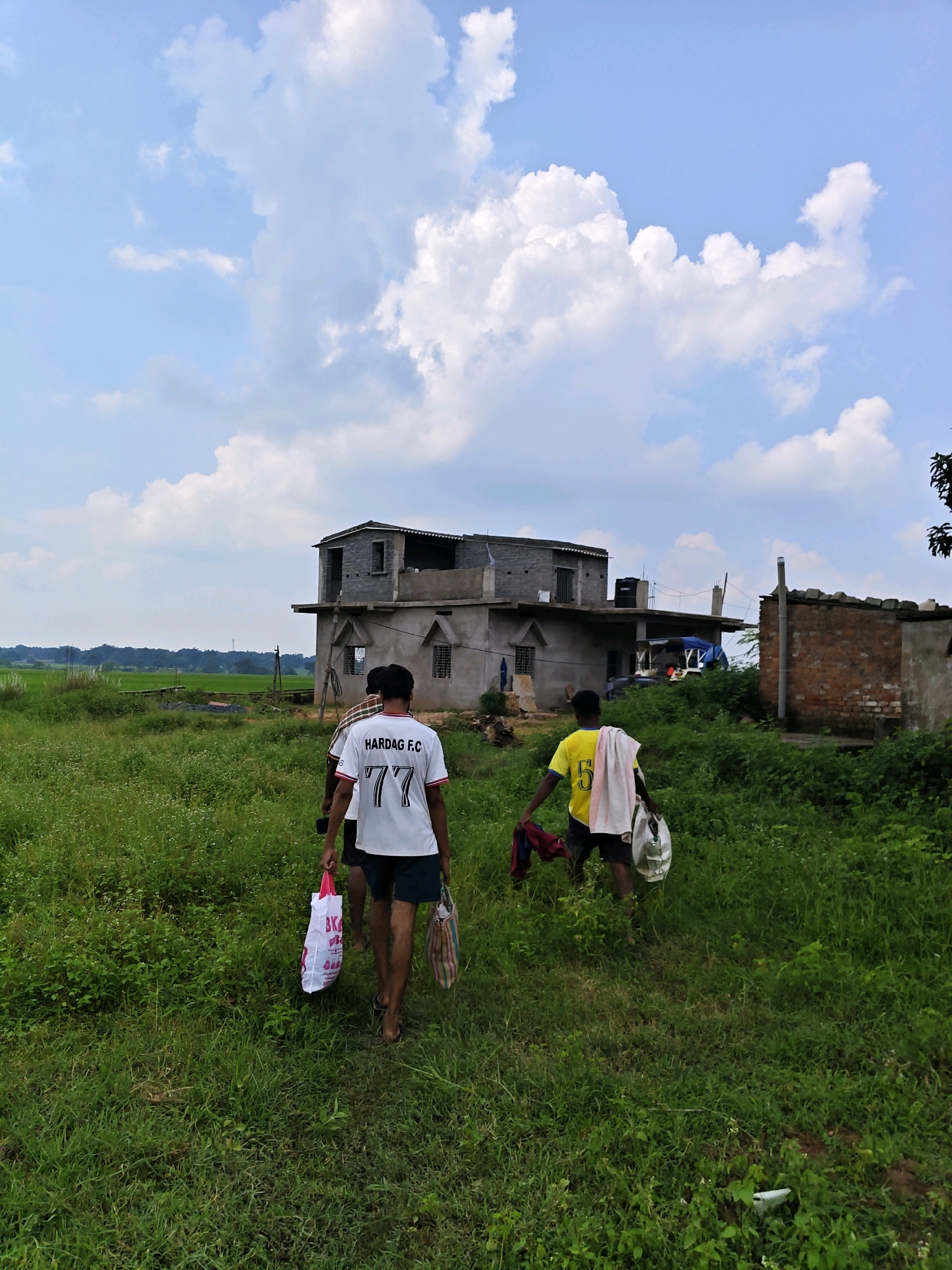 The image shows three young males walking through a grassy field in Sosai, India. The sky is a bright blue with scattered white clouds. In the background, there is a partially constructed building with multiple levels and a water tank on the roof. To the right of the building, there are structures made of brick and what appears to be corrugated metal. The male in the foreground on the left is wearing a white t-shirt with "HARDAG F.C. 77" printed on the back and dark shorts. He is carrying a white plastic bag in his left hand and a striped bag in his right. The male in the middle is partially visible and is wearing a checkered shirt. The male on the right is wearing a yellow t-shirt with the number "5" on it and dark shorts. He is carrying a white towel over his shoulder and a white plastic bag in his right hand, with some red clothing in his left hand. The environment is rural, with lush green grass and vegetation.