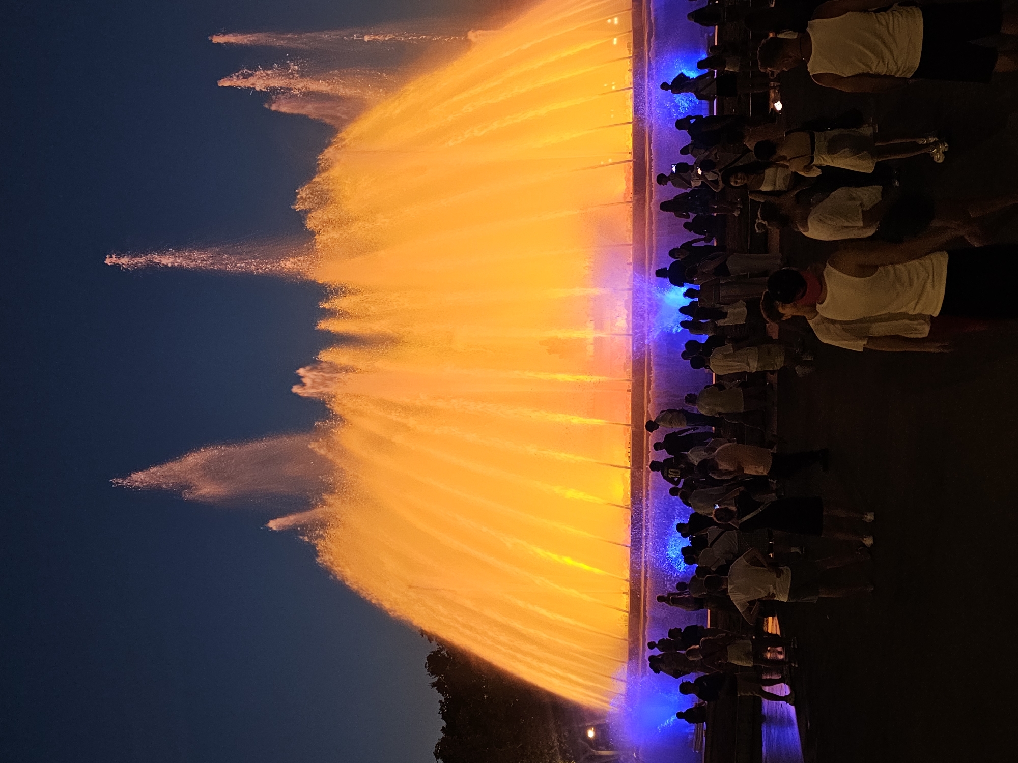 The image captures a night view of the Magic Fountain of Montjuïc in Barcelona, Spain. A large, illuminated fountain is the central subject, with cascading water lit in vibrant shades of orange and yellow, creating a dramatic effect against the dark blue night sky. Blue lights are visible at the base of the fountain. A crowd of people, silhouetted against the bright fountain, stands in the foreground, observing the display. The people are of varying ages and are dressed in casual attire. Some are standing, while others appear to be walking or mingling. The overall scene conveys a sense of public gathering and visual spectacle, typical of an evening attraction in a city.