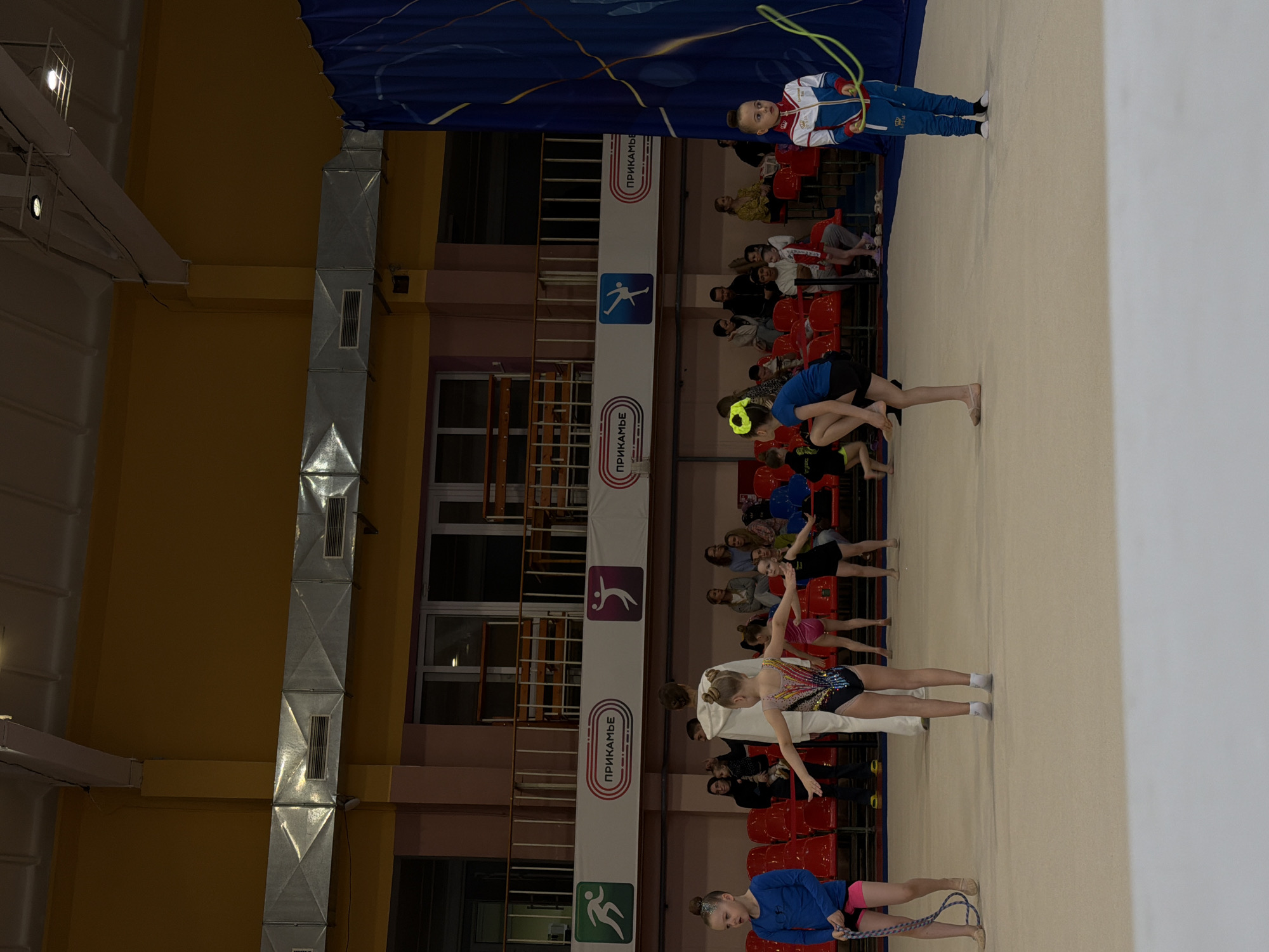 The image shows a group of young gymnasts in a gymnasium, likely during a training session or competition. Gymnasts are seen in various poses, including stretching and possibly performing routines. Some are in leotards, while others wear athletic wear. Spectators, likely parents or coaches, are seated in red chairs along the sidelines. The gymnasium has an indoor layout with windows, ventilation ducts, and banners with the word "ПРИКАМЬЕ" (Prikamye) and stylized athlete figures. The banners suggest a connection to the Prikamye region. The setting appears to be in Perm, Russia, given the Russian word on the banners, and that Prikamye is a region in the Perm Krai of Russia. One gymnast holds a jump rope.
