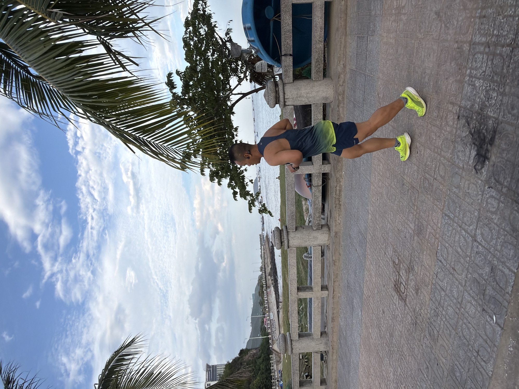 A man is jogging on a paved pathway along the coastline in Nha Trang, Vietnam. The man is wearing a dark blue and yellow tank top and dark blue shorts, with bright yellow running shoes. He has short hair and is wearing earbuds. The pathway is made of square, patterned tiles. Behind the pathway is a low, stone-and-concrete wall with decorative posts. Beyond the wall, there is a grassy area with a few small boats overturned on it, and a large blue boat. A tree with green leaves is visible to the right of the runner. In the background, the ocean stretches to the horizon under a partly cloudy sky. Buildings and a hillside are visible on the left side of the image, with palm trees in the foreground. The scene appears to be set during the daytime, likely late afternoon or early evening.