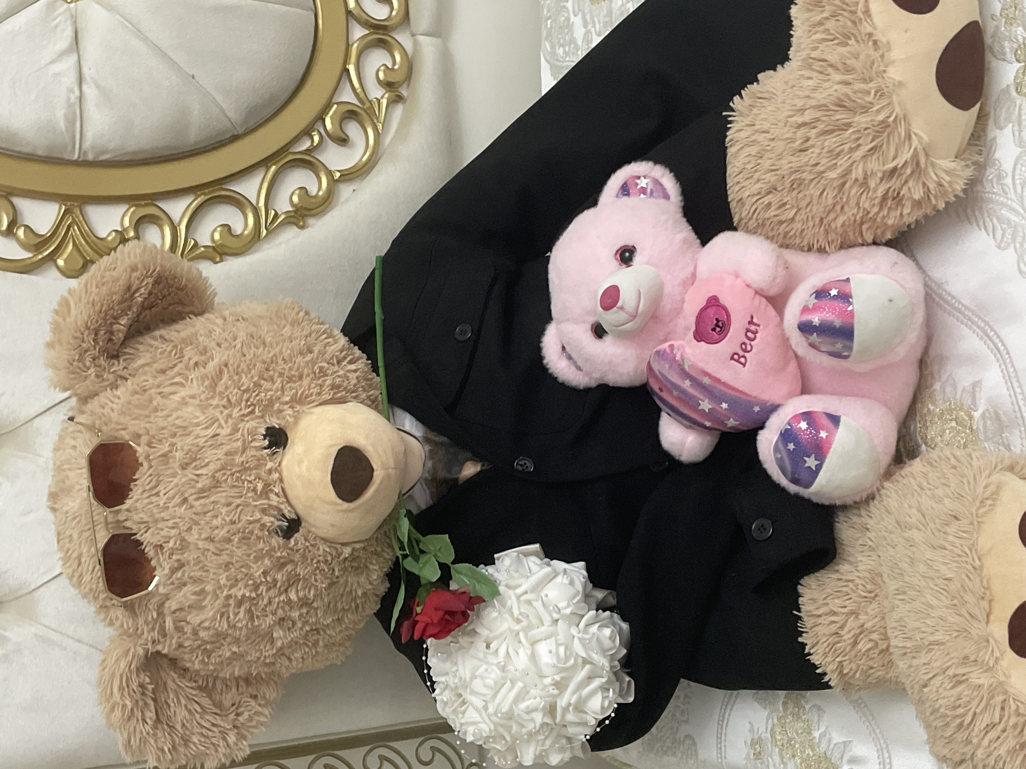 The image presents a charming indoor arrangement centered around teddy bears and floral decorations. In the foreground, a large, light brown teddy bear lies on its back with hexagonal, gold-framed sunglasses resting on its head. Adjacent to it, a black jacket or suit forms a base for a smaller, pink teddy bear. This pink bear clutches a heart-shaped pillow with the word "Bear" written on it and features glittery, star-patterned fabric on its inner ears and paws.
Nestled between the two primary bears on the black jacket is a bouquet of white artificial roses adorned with small pearl accents, accompanied by a single red rose with a green stem. Additional light brown teddy bear parts are visible in the upper and lower right corners of the frame, hinting at more plush toys in the scene. The setting includes white quilted upholstery, possibly a headboard or cushion, and a white fabric with a subtle floral pattern. A decorative, ornate gold frame is prominently visible on the left side, suggesting an elegant interior. The composition evokes a sweet and playful, possibly romantic, theme in what appears to be a room within Ihtiman, Bulgaria.