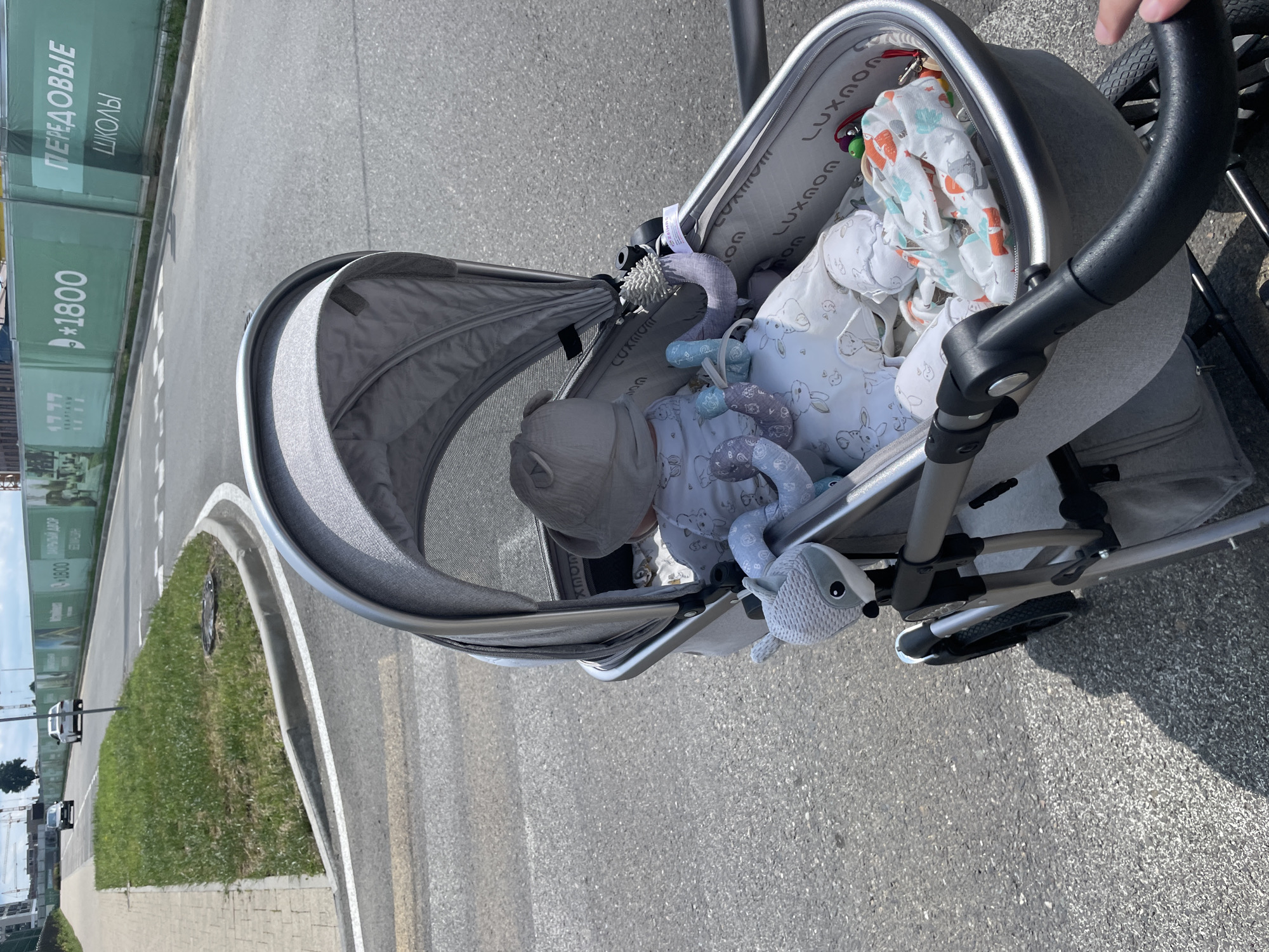 A baby in a stroller is being pushed on a paved road in Stavropol, Russia. The stroller is gray and has a canopy. The baby is wearing a hat and a onesie with a rabbit pattern. There are toys attached to the stroller. In the background, there is a green fence with advertisements for "PEREDOVYE SHKOLY" which translates to "ADVANCED SCHOOLS." Cars are visible on the road, and there is a sidewalk with grass next to it. The sky is cloudy.