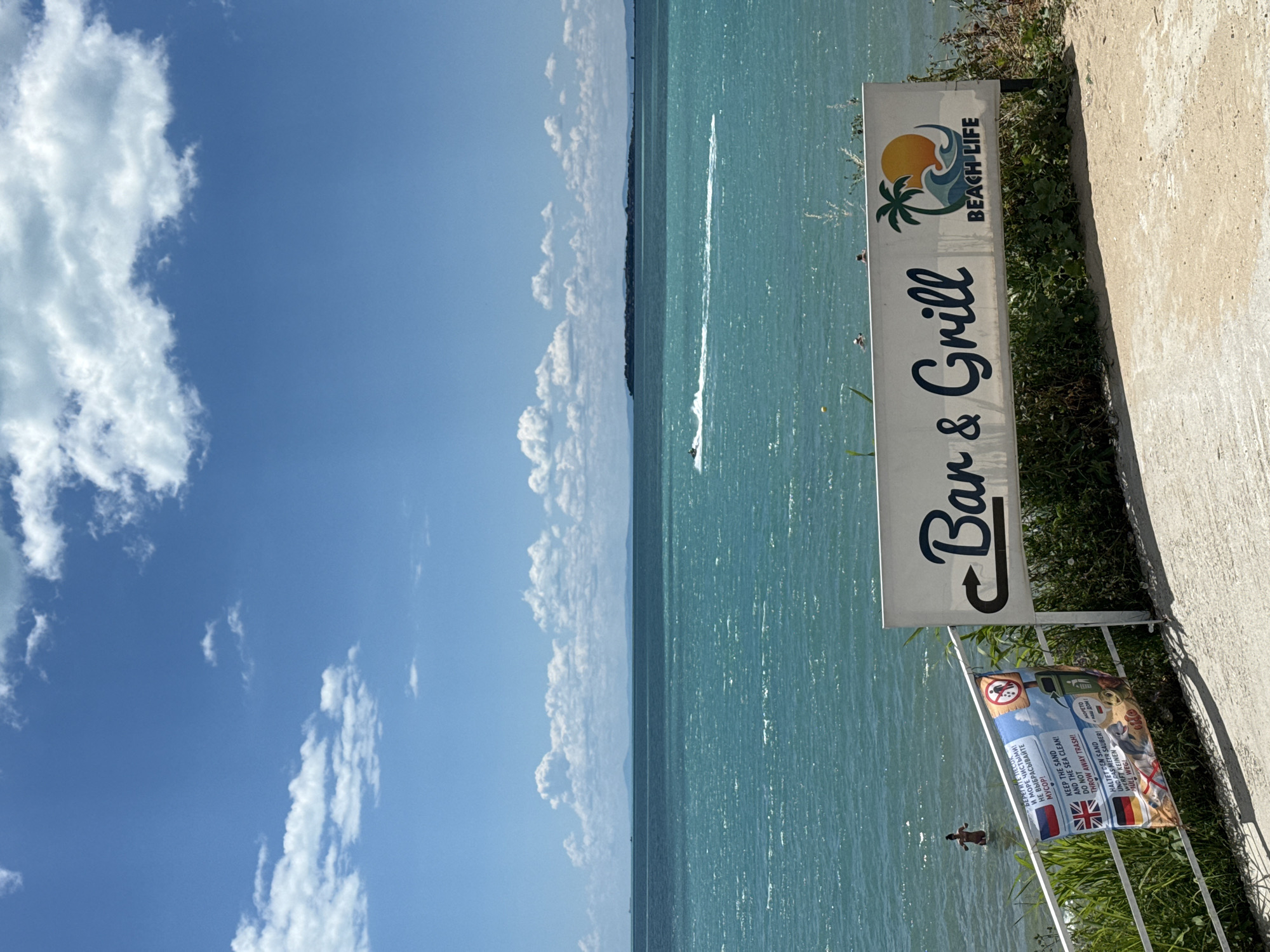 The image depicts a sunny day at a beach in Sveti Vlas, Bulgaria. In the foreground, a white sign with dark blue lettering reads "Bar & Grill" and features a graphic of a palm tree against a sunset over waves, with the text "BEACH LIFE" below. Adjacent to the sign, a smaller banner displays flags of Russia, the UK, and Germany, along with text in Cyrillic, English, and German, conveying a message about keeping the sand and sea clean and not throwing away trash. A metal railing runs along the edge of the scene, adorned with some green foliage. In the background, the turquoise sea stretches towards the horizon, dotted with a few distant islands. A jet ski can be seen creating a white wake on the water, and several people are visible in the sea, with one person standing waist-deep in the water. The sky is blue with scattered white clouds.