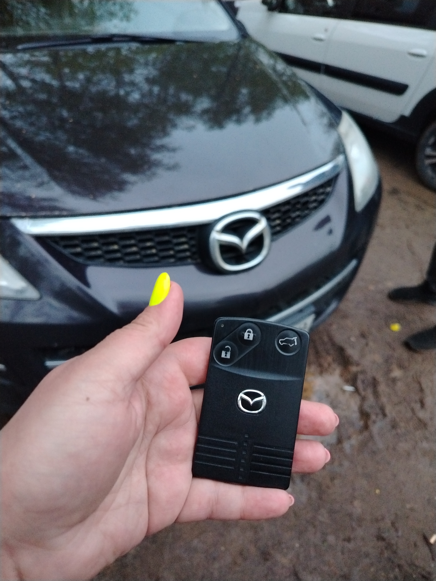 A close-up, eye-level shot shows a hand holding a black Mazda car key in front of a dark-colored Mazda car. The hand has a bright yellow thumbnail. The Mazda key has the Mazda logo and three buttons: two lock buttons and a trunk release button. The Mazda car is partially visible, showing the grill and headlights. A second white SUV-style vehicle is also partially visible in the background. The ground appears to be wet or muddy. A person wearing black shoes is standing on the right side of the frame. The location is in the city of Chaikovsky, Russia.