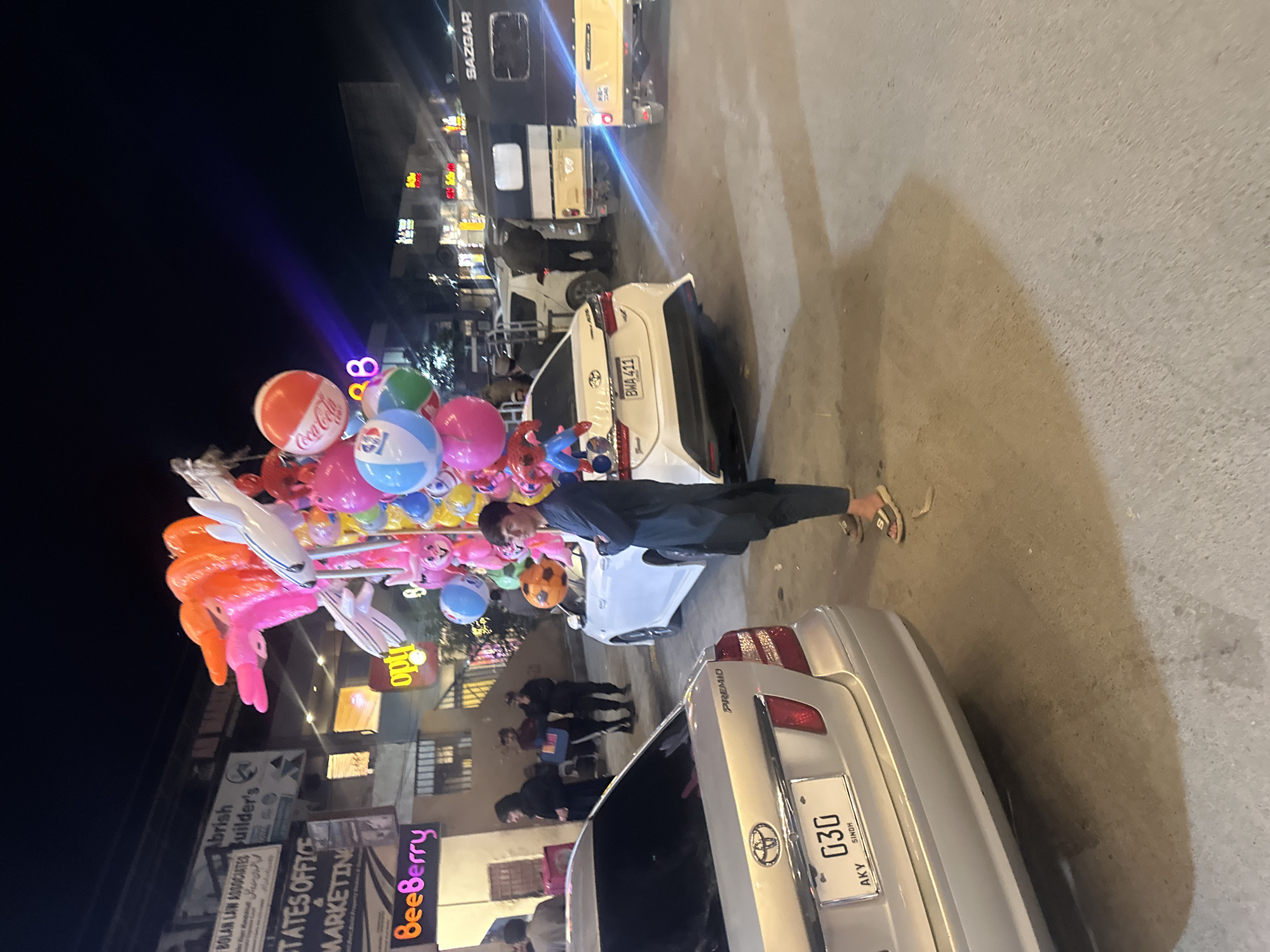 The image depicts a nighttime street scene in Quetta City Tehsil, Pakistan. Dominating the frame is a balloon vendor holding a large cluster of balloons, including recognizable Coca-Cola and Pepsi-branded ones. The vendor is standing amidst parked vehicles, primarily Toyota cars. The vehicles are oriented in different directions. The street is lined with buildings, some displaying lit signage, including "Beeberry", "STATES OFFICE MARKETING", "BOLAN LAW ASSOCIATES Builder's", and "opty". There are people walking in the background. In the far background, a "SAZGAR"-branded vehicle is visible. The environment is urban and the setting is at night.