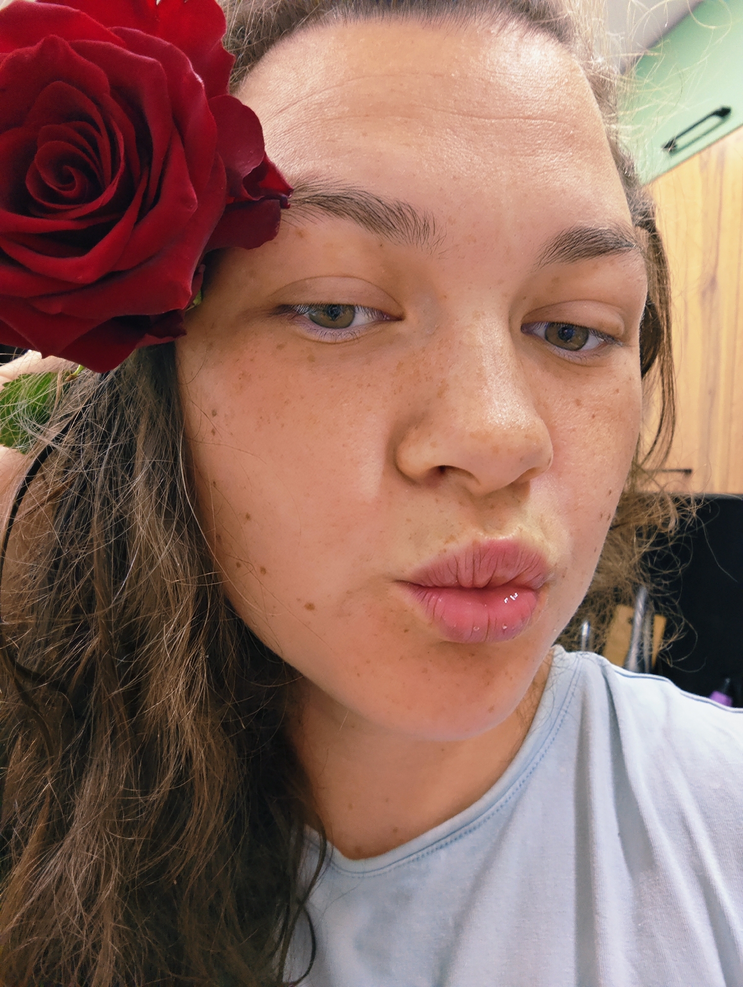 A close-up captures a young woman with fair skin and numerous freckles, a large red rose positioned near her left temple. Her hair is long and wavy, a mix of brown and blonde strands. She has light brown eyes and is making a kissing face with her lips slightly pursed. She is wearing a light blue t-shirt. The background is indistinct, appearing to show a cabinet with a light brown wooden surface. The specific location is not identifiable within this image.