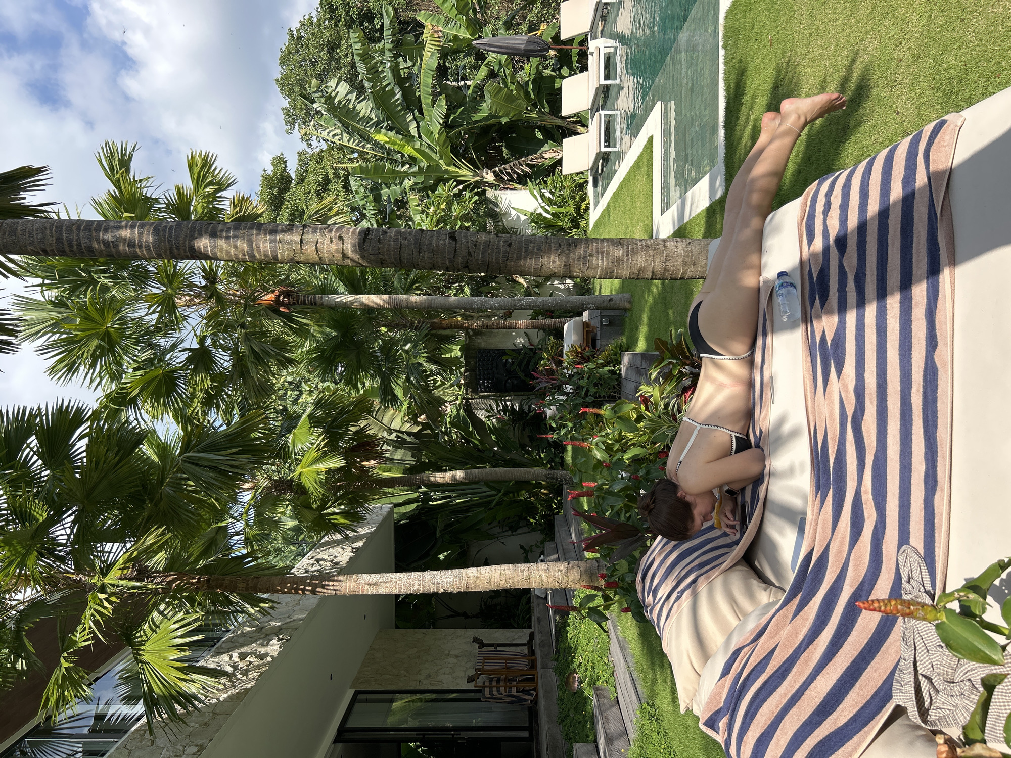 A woman in a black and white bikini sunbathes on a striped lounge chair in a lush, tropical garden. She is lying on her stomach, looking down at her phone, with her head resting on her hands. Behind her, a swimming pool with lounge chairs and umbrellas is visible, as well as a modern house with stone accents and large windows. Palm trees and other dense green foliage surround the area, suggesting a warm, sunny climate. The scene appears to be set in a resort or a private villa, possibly in a location known for its tropical beauty.