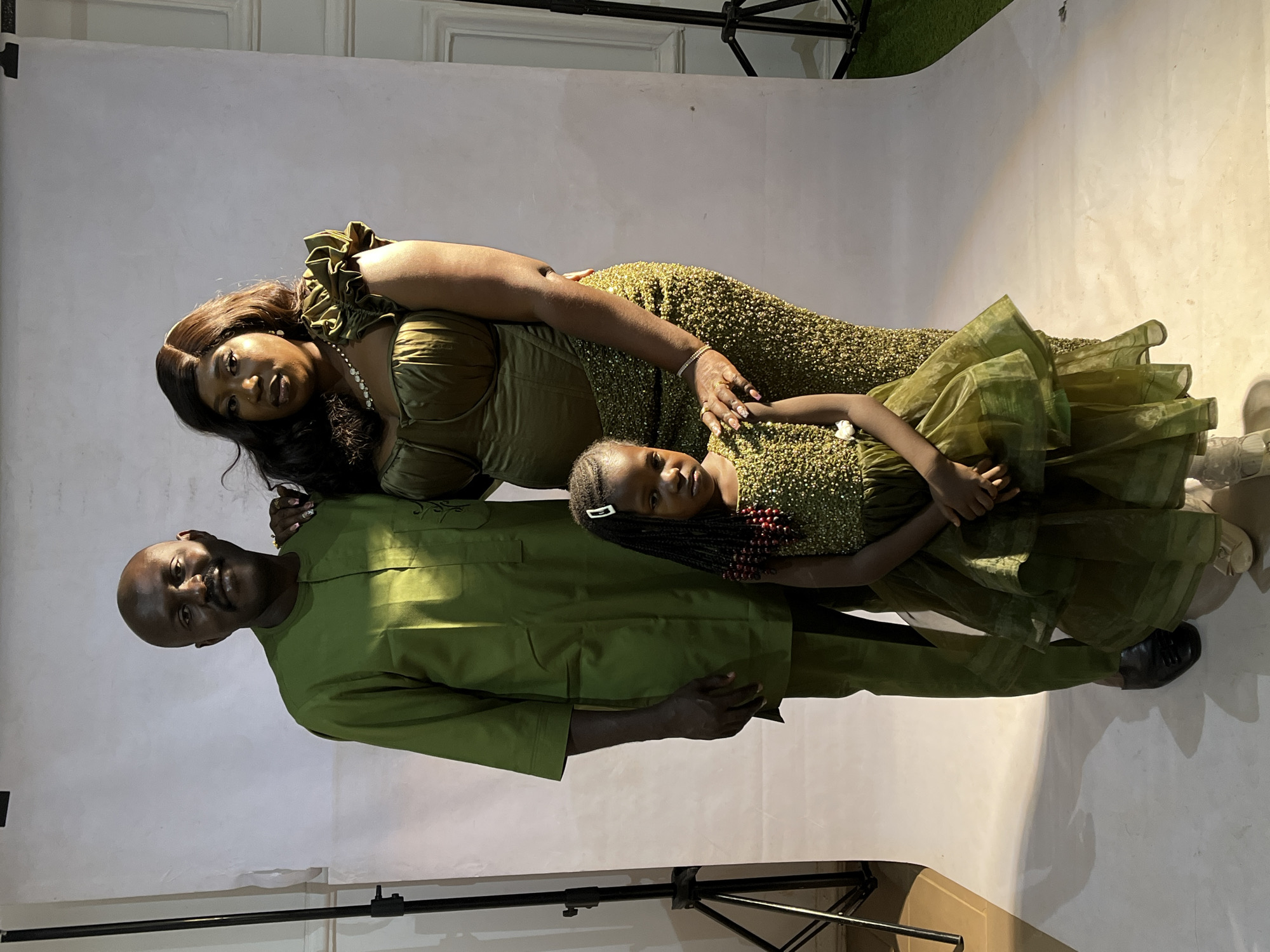 A family portrait featuring three people is taken in a studio. The studio has a plain white backdrop. A lighting rig and what appears to be a patch of artificial grass are visible in the background. The family consists of a man, a woman, and a young girl.
The man is bald, wearing a light olive-green long-sleeved tunic and trousers. He is positioned on the left side of the frame, supporting the woman.
The woman is wearing a sparkling dress, olive-green, with shoulder ruffles and a beaded necklace. Her hair is styled in long dark waves. She is lying down, supported by the man.
The young girl is wearing a dress matching the color scheme, with an olive-green shimmering top and an organza skirt. She is lying between the man and woman. She is wearing a beaded head accessory.
The family appears to be posing for a studio portrait.
It's unlikely that the location in the city of Jos, Nigeria, can be determined from the photograph.