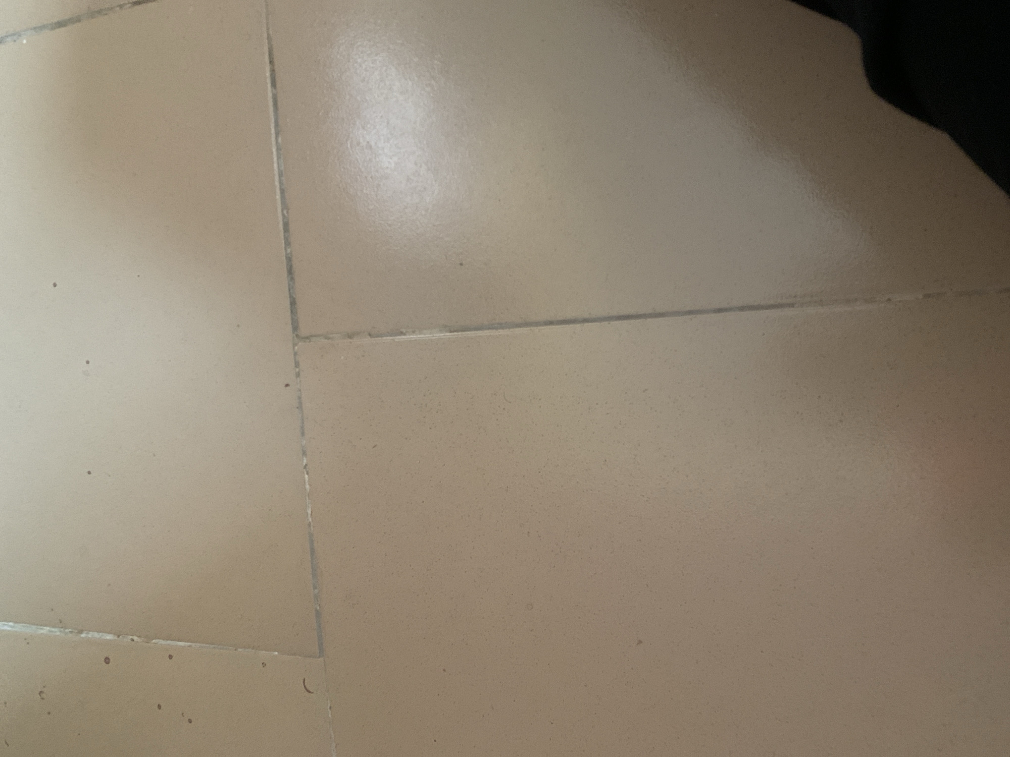 The image shows a close-up, overhead view of a tiled floor. The tiles are a uniform, light beige color and appear to be made of ceramic or a similar material. They are laid in a grid pattern with visible grout lines between them. The floor shows signs of wear and tear, with some minor scuff marks and a few small dark specks scattered across the surface. The lighting in the image is somewhat uneven, creating subtle shadows and highlights that emphasize the texture of the tiles. In the bottom right corner of the image, a portion of a dark, indistinct object, possibly clothing or a bag, is visible. The overall impression is of a functional, everyday floor. The location in Jauro Yinu, Nigeria, is not visually represented in the image itself.