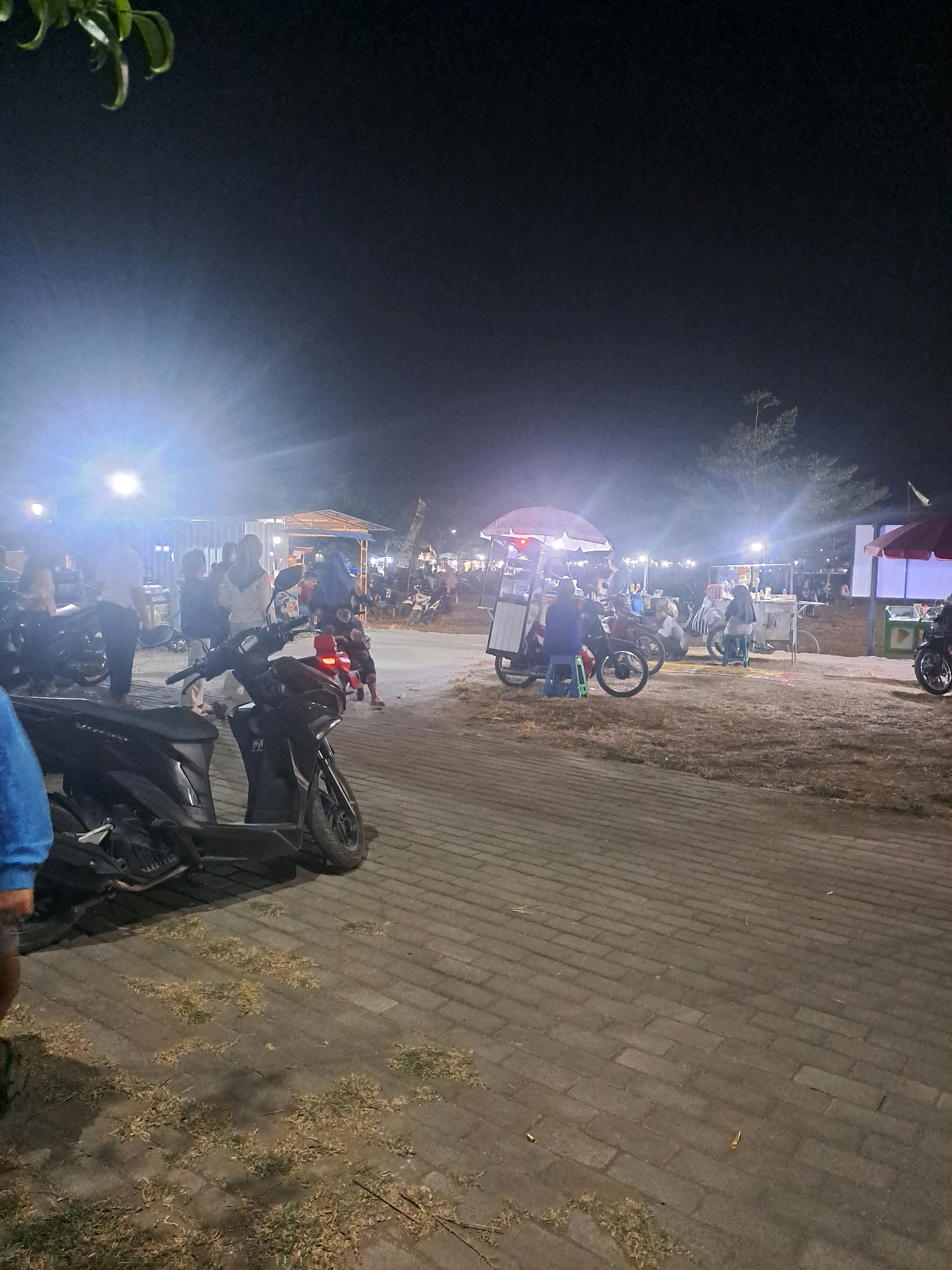 The image depicts a nighttime scene at a street market or food stall area, likely in Sumberejo, Indonesia, given the context provided. A paved walkway with scattered dry grass and debris is in the foreground. Several parked motorcycles, primarily scooters, are visible, with one dark-colored scooter in sharp focus in the lower left portion of the frame. In the mid-ground, various food stalls are set up, some with umbrellas providing cover. People are seen gathered around these stalls, some appearing to be customers and others vendors. Bright lights illuminate the scene from various sources, including the stalls and possibly streetlights, creating glare. Trees are visible in the background, silhouetted against the night sky. The atmosphere suggests a bustling evening market environment.