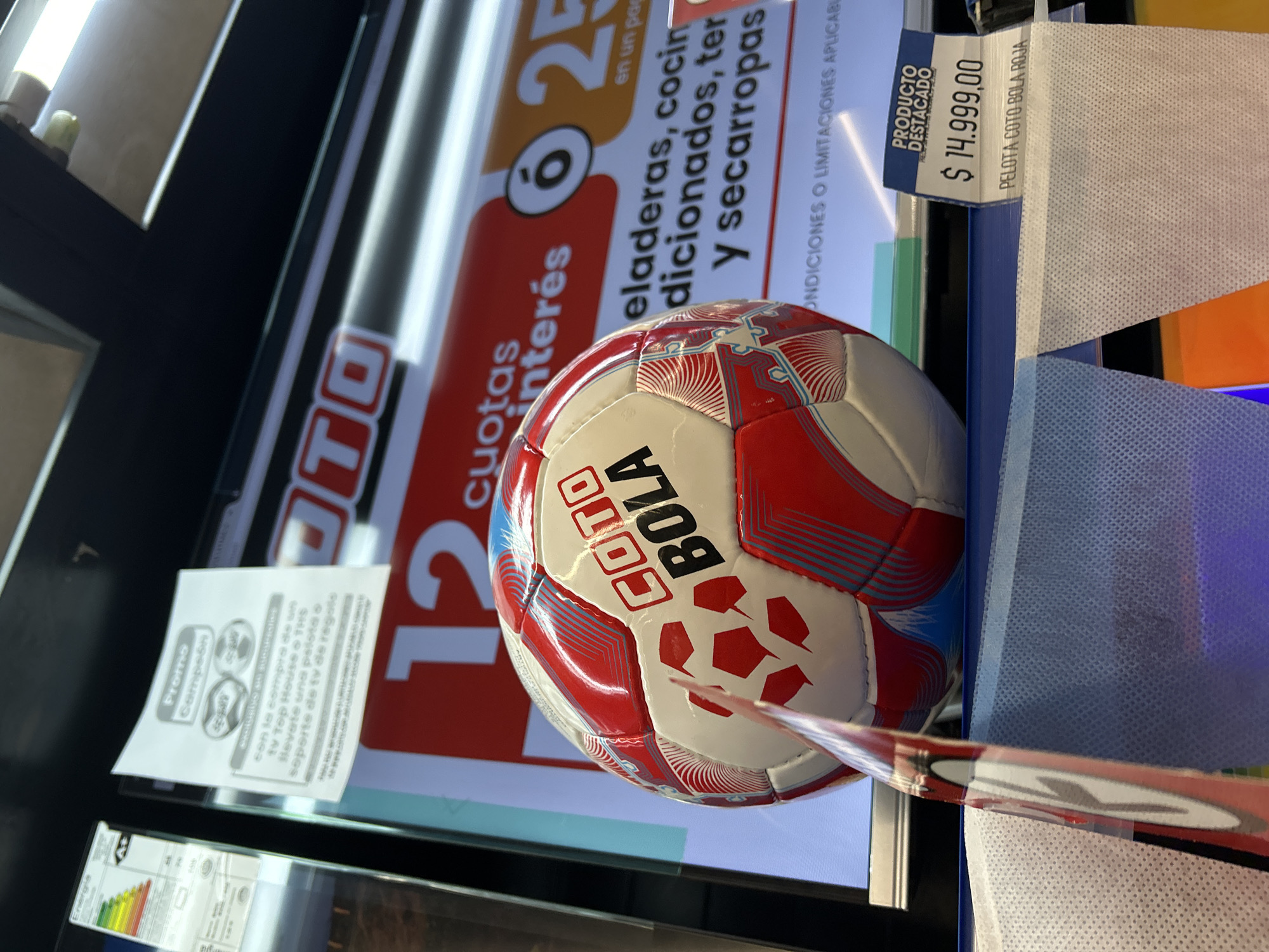 The image depicts a COTO Bola soccer ball priced at $14,999.00 Argentine pesos on display within a COTO supermarket environment. The COTO logo and promotional material related to the purchase of TVs for a complimentary soccer ball or TV stand is also visible. The sign displays "12 cuotas interes" (12 interest-free installments) and mentions "heladeras, cocinas" (refrigerators, cookers), suggesting a section within the supermarket focusing on home appliances.