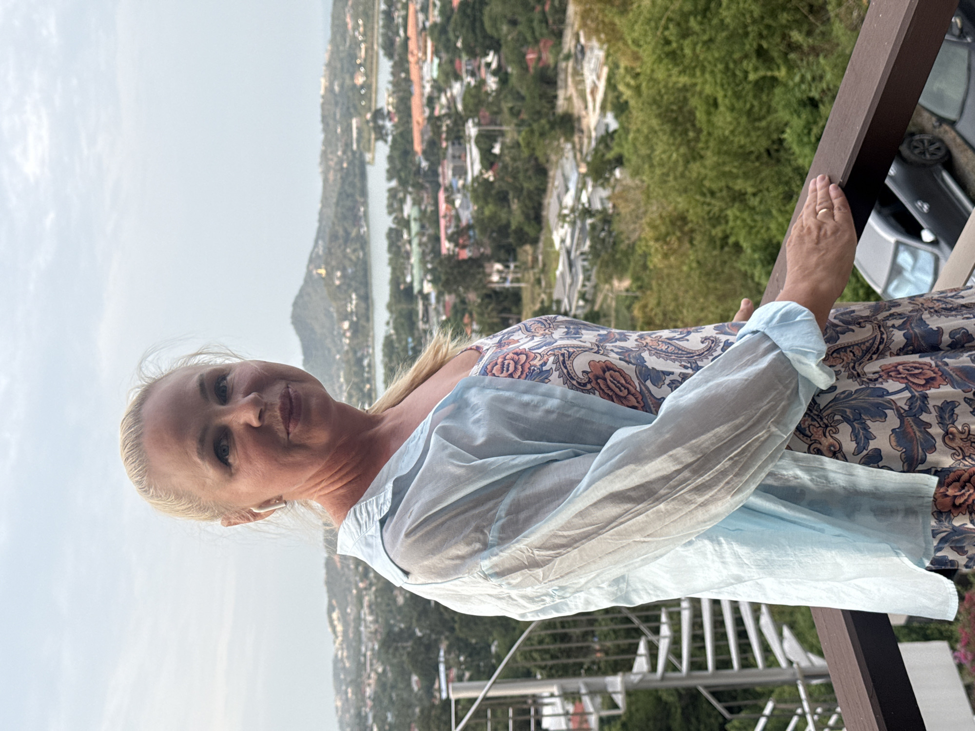 A woman with blonde hair pulled back stands on a balcony overlooking a city and a body of water. She is wearing a light blue unbuttoned shirt over a floral print dress. She has on white wireless earbuds. Her right hand rests on the wooden railing of the balcony. In the background, there are buildings, lush green trees, a hill with a golden structure on top, and a body of water. A spiral staircase is visible to the left of the woman. Below the balcony, two cars are parked. The sky is overcast. The scene is likely in the Ko Samui District of Thailand, given the architecture and landscape.