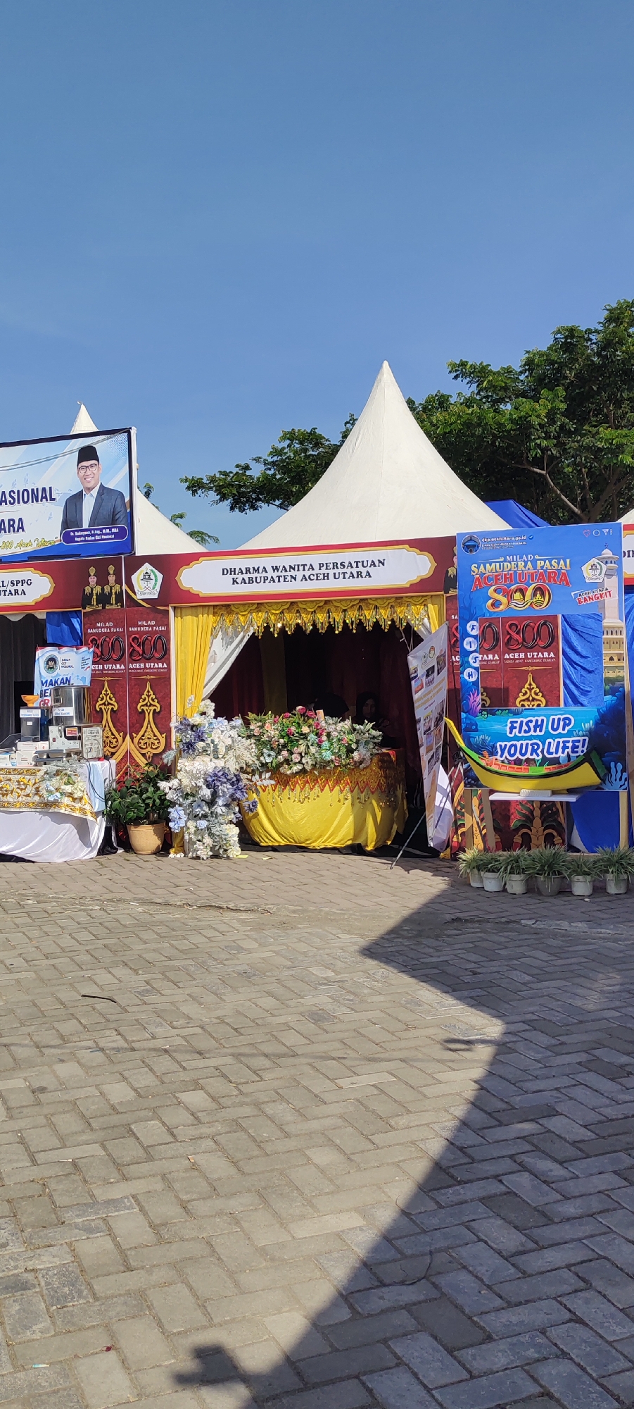 The image depicts an outdoor event, possibly a festival or exhibition, set against a clear blue sky and a backdrop of trees. In the foreground, a paved walkway made of grey interlocking bricks is visible, with shadows cast upon it.
The central focus is a large, white tent with a pointed peak. In front of the tent, a display is set up. To the left of the tent entrance, there's a table covered with a white tablecloth, holding various items that appear to be food or products, along with a coffee maker or similar appliance. Next to this table, a potted plant sits on the paved ground.
The entrance to the tent is adorned with yellow and red drapes, with decorative golden trim. Inside the tent, a table is covered with a yellow cloth and decorated with an abundant floral arrangement in shades of pink, white, and blue. Behind the table, a person wearing a dark headscarf is partially visible, suggesting they are attending to the display.
To the right of the tent entrance, a vertical banner with the text "SAMUDERA PASAI ACEH UTARA 800" is prominently displayed. Below this, another banner reads "FISH UP YOUR LIFE!". In front of this banner, a row of small potted plants lines the paved area.
Further to the left of the main tent, another tent structure is partially visible, featuring a large banner with a picture of a man in formal attire and text including "NASIONAL" and "ACEH UTARA". Another smaller banner to the left of this shows three figures in uniform and text.
The overall impression is one of a well-organized outdoor event with various displays and stalls. The presence of banners with place names like "ACEH UTARA" and Indonesian text suggests the location is likely in Indonesia, specifically in or near Meunasahaluedrienlhoksukon Barat, as indicated. The weather appears to be bright and sunny, conducive to an outdoor gathering.
