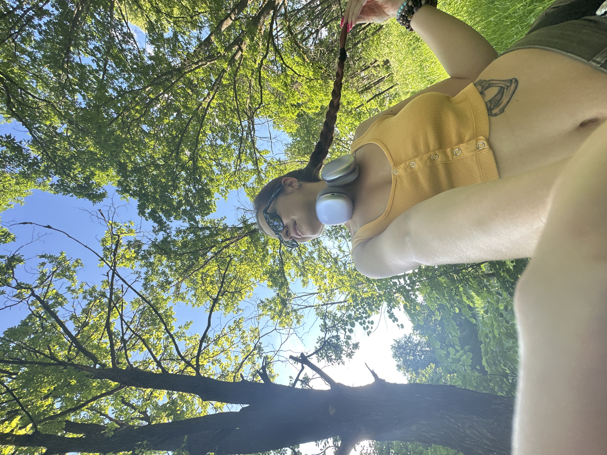 The image shows a woman with braided brown hair, wearing a yellow tank top and sunglasses. She has headphones around her neck and is holding her braid with her right hand, which has pink fingernails and is adorned with bracelets. She has a tattoo on her stomach. The woman is outdoors, surrounded by trees and grass, under a clear blue sky. The lighting suggests it is daytime, possibly afternoon, with the sun filtering through the leaves. The background shows a lush green environment with tall trees and dense foliage. The perspective suggests the photo was taken from a low angle, possibly by the woman herself. The location is specified as the city of Vladimir, Russia.